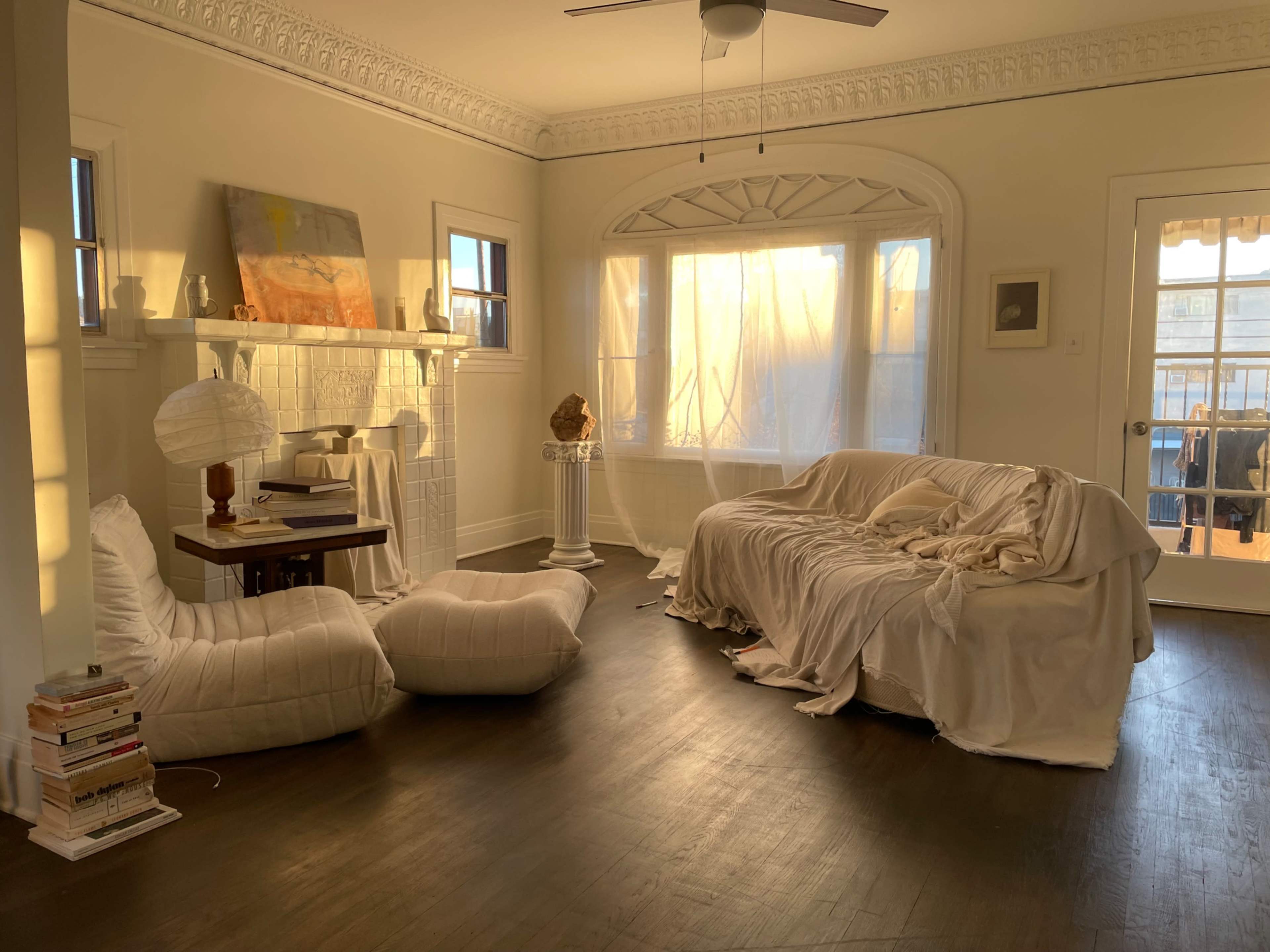 The sunlight streams through sheer curtains into a spacious living room with a couch draped in white fabric, a small table, and two large floor cushions.
