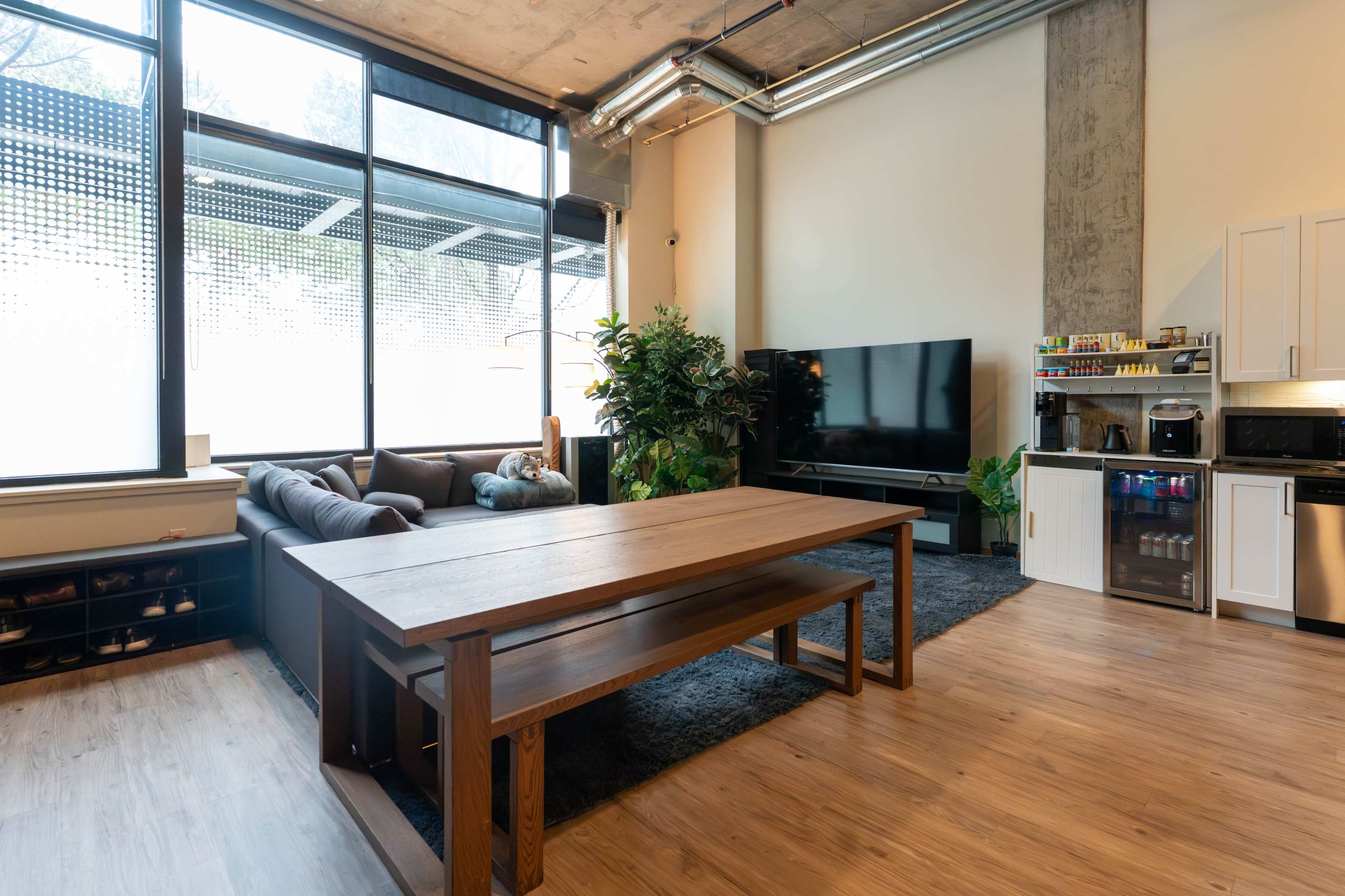 The image shows a modern living space featuring a large wooden dining table, a dark couch, a television, and a kitchenette in a well-lit room with large windows.