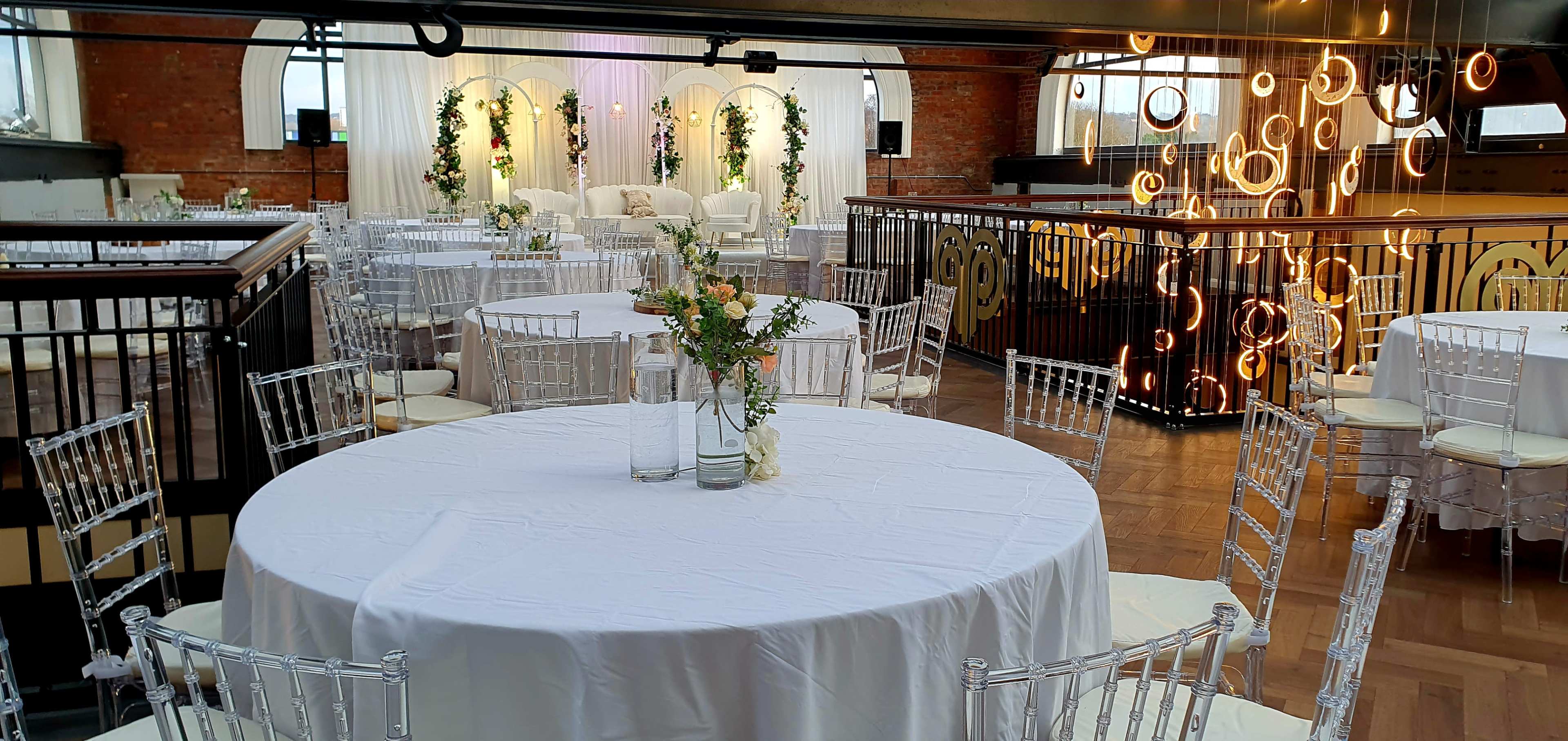 The image shows a decorated event space with circular tables set up and a stage area featuring floral arrangements and seating in the background.