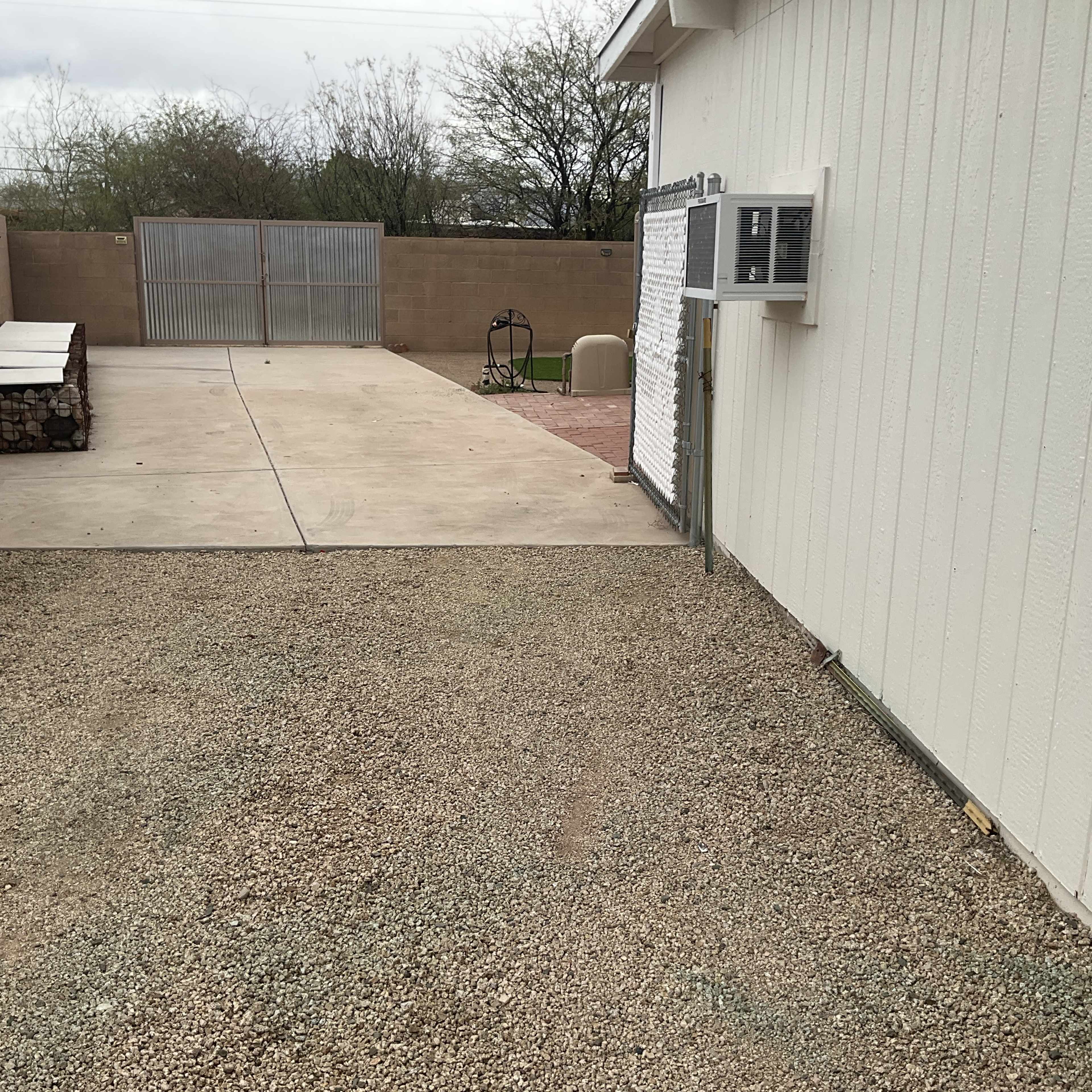 The image shows a gravel area next to a concrete patio and a fence with a metal gate in an outdoor setting.