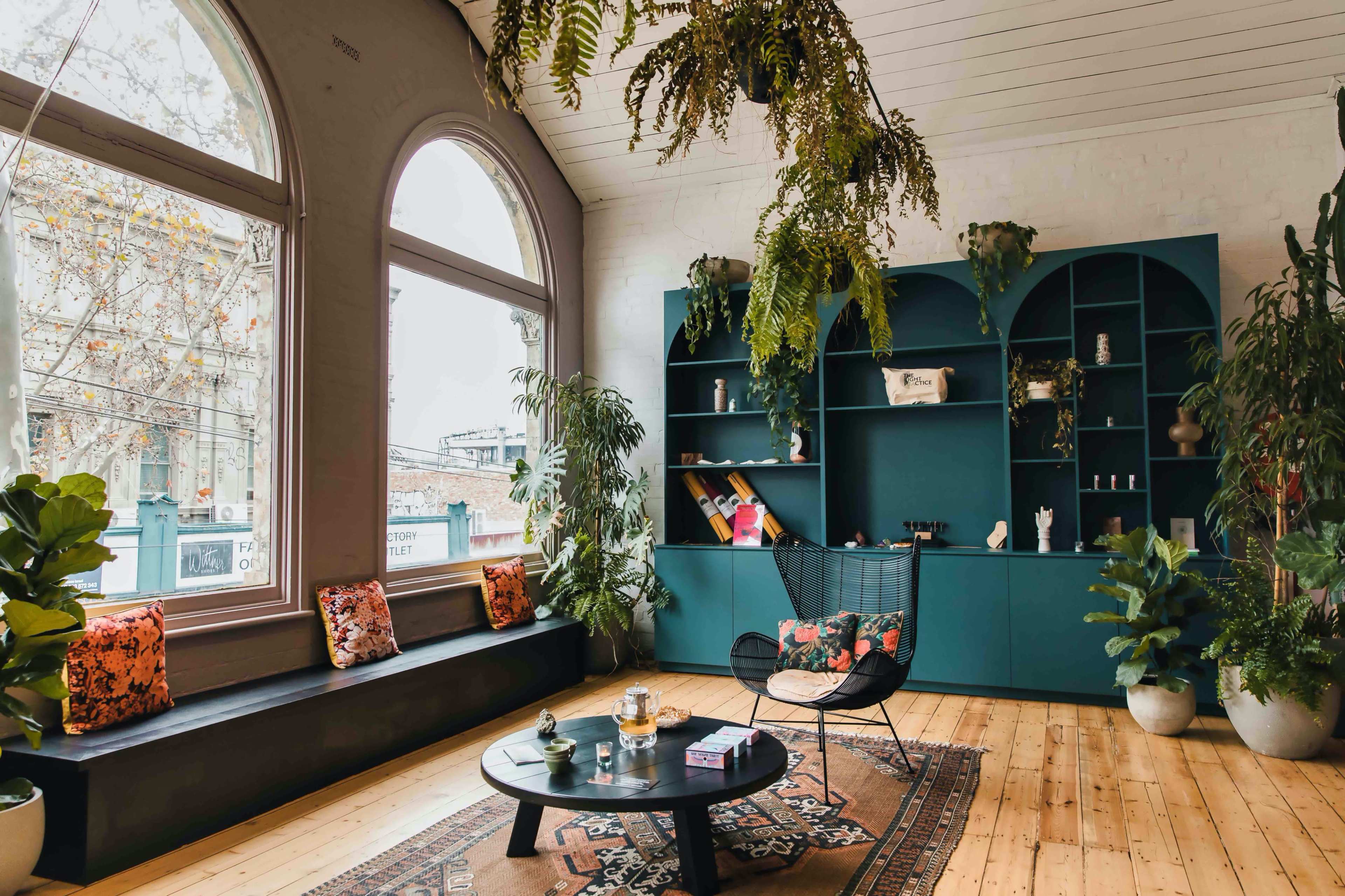 The room features large arched windows, a dark green shelving unit, a circular coffee table, and various potted plants.