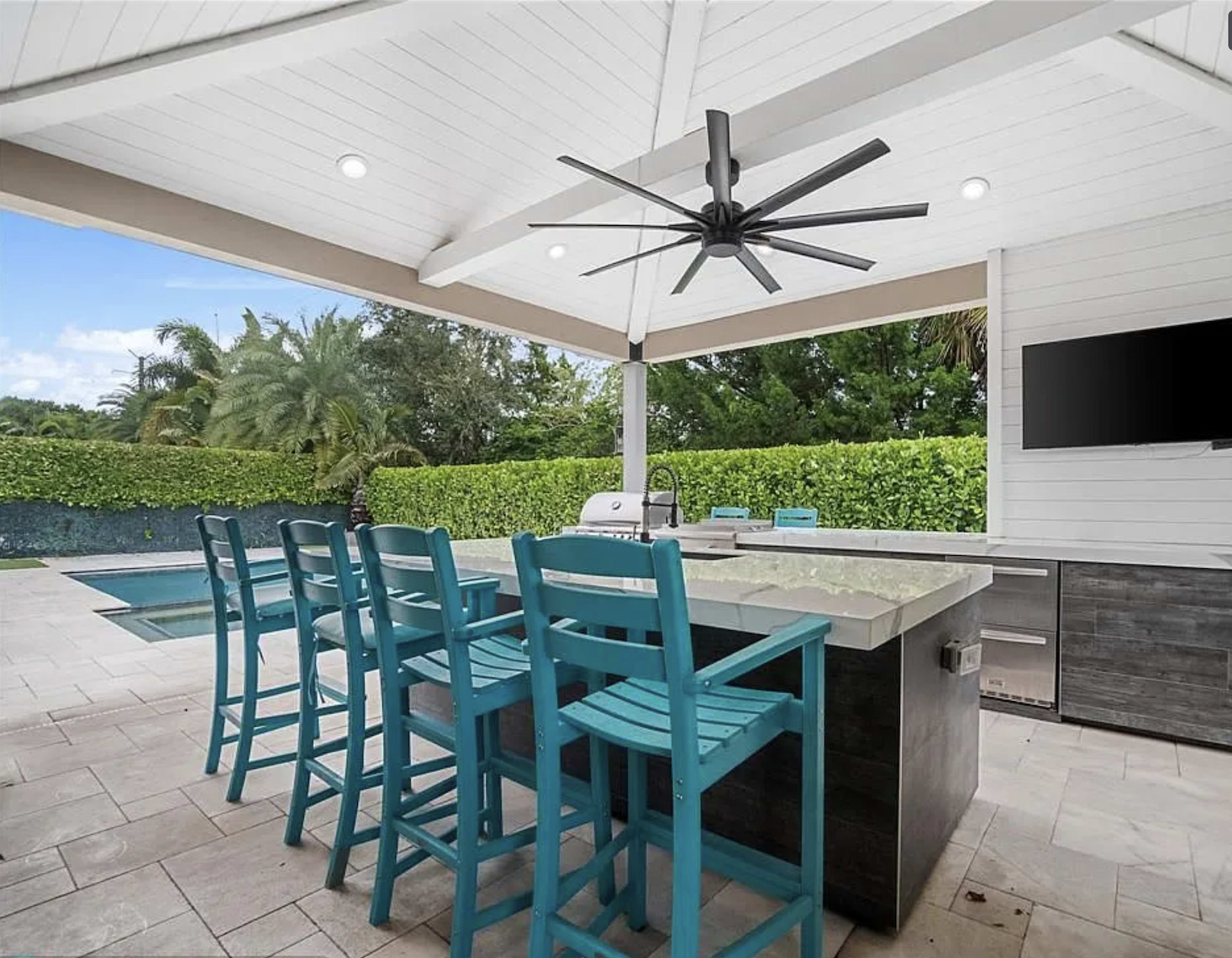 The image shows an outdoor kitchen area with a bar, featuring turquoise stools, a grill, and a swimming pool in the background.