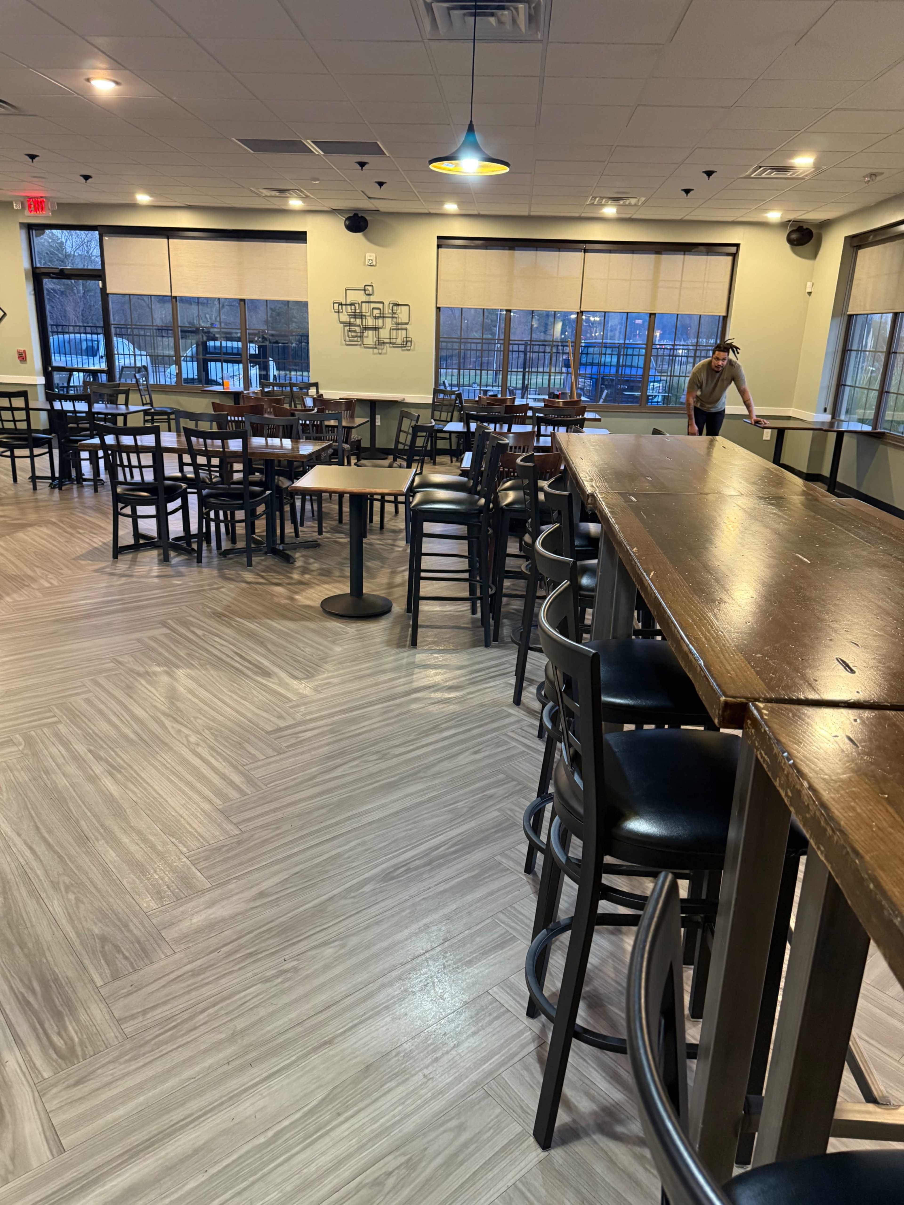 The image shows an empty restaurant interior with wooden tables and black chairs, and a staff member cleaning a table in the background.