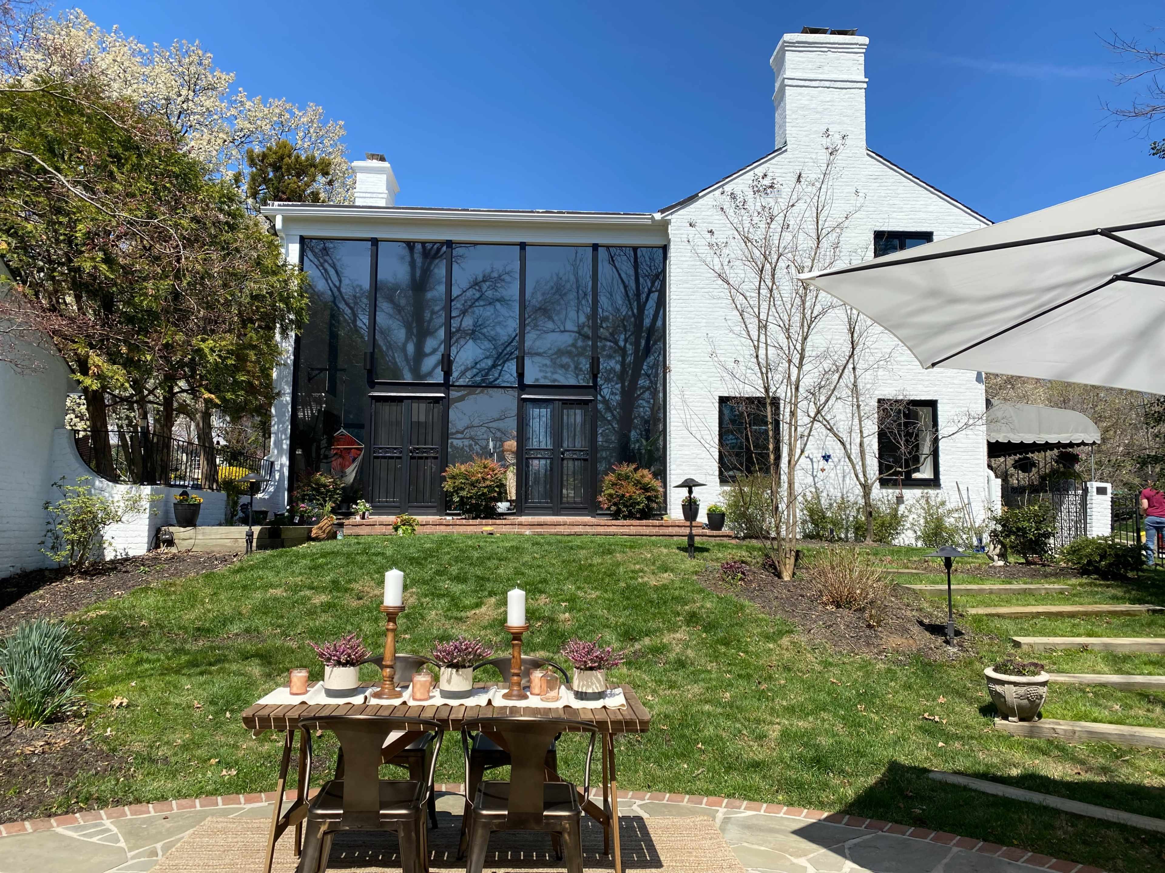 The image shows a modern white house with large black-framed windows, a green lawn in front, and a wooden table set with candles and decorative items.