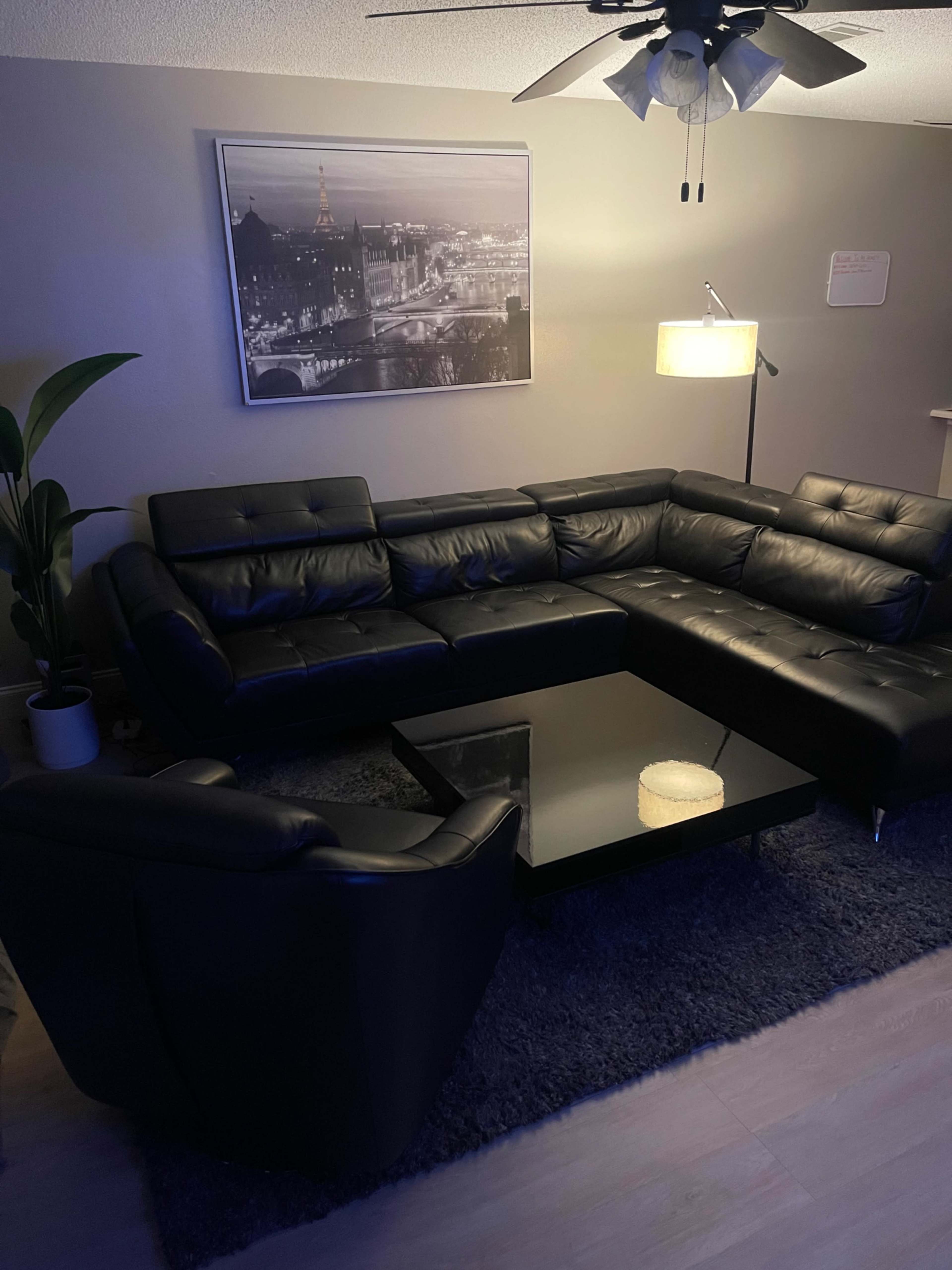 A modern living room featuring a black leather sectional sofa, a glass coffee table, and a floor lamp, with a large framed black and white photograph on the wall.