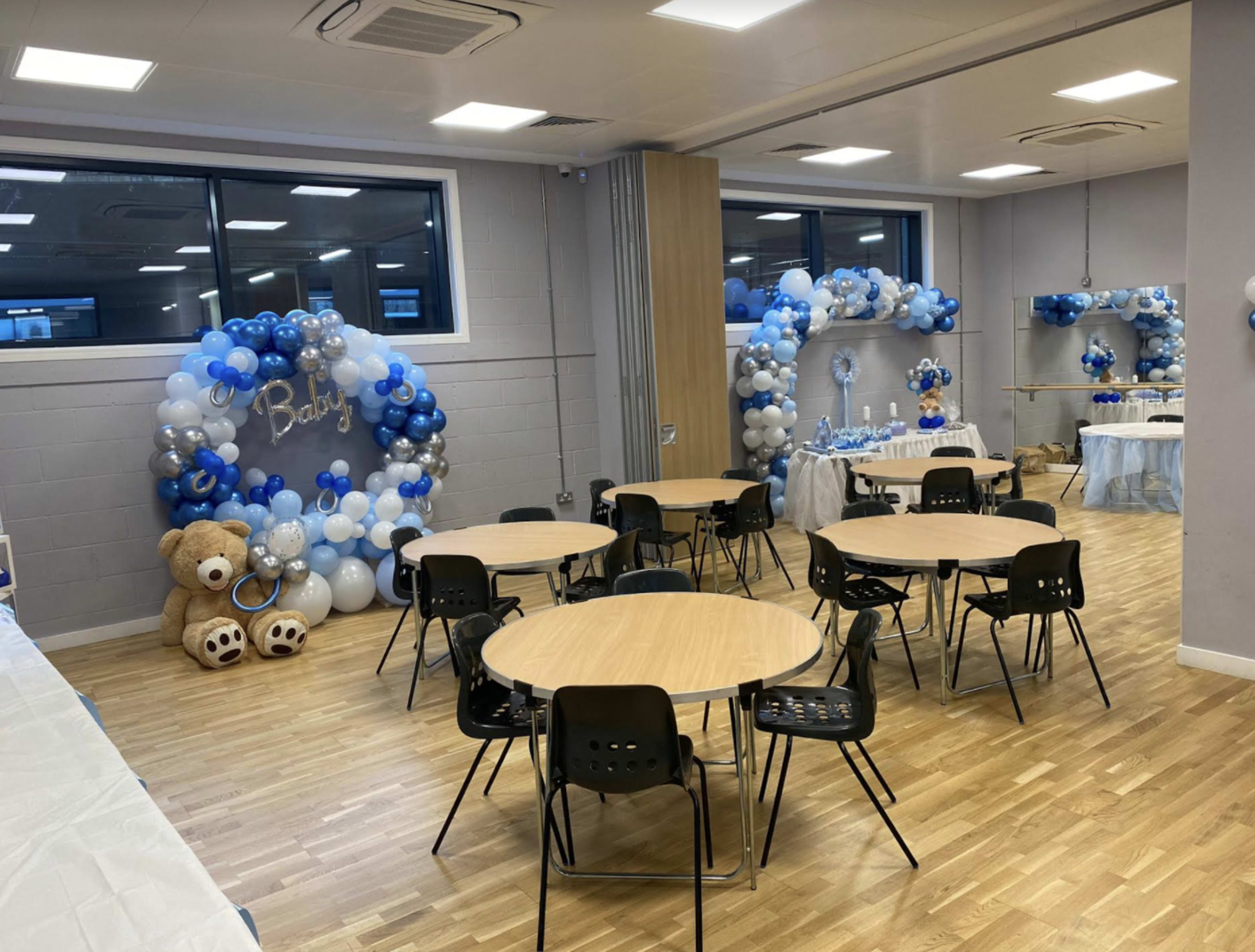 A decorated event space featuring balloon arches in shades of blue, tables with black chairs, and teddy bear centerpieces.
