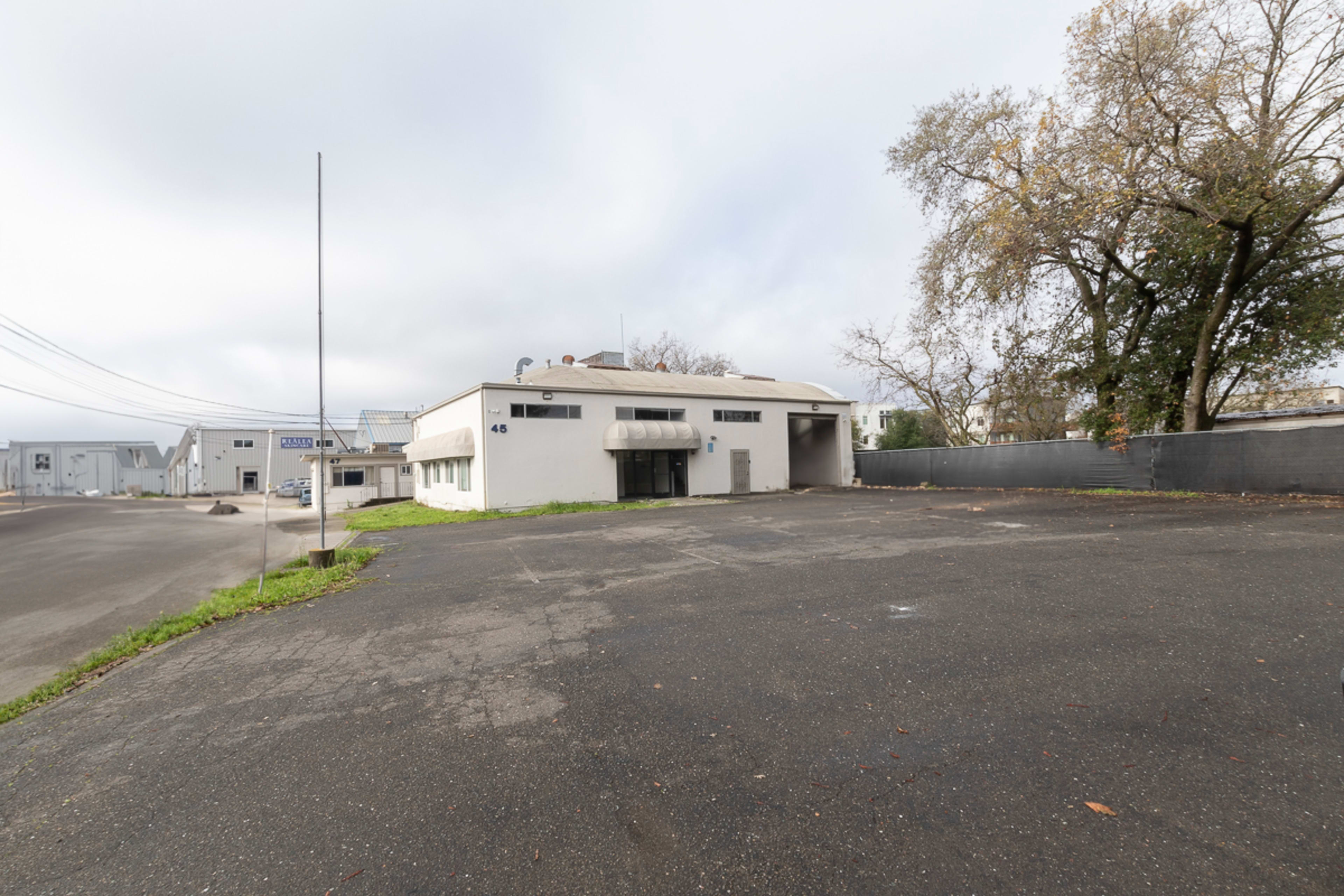 An empty industrial parking lot in front of a white commercial building partially surrounded by trees and utility poles.