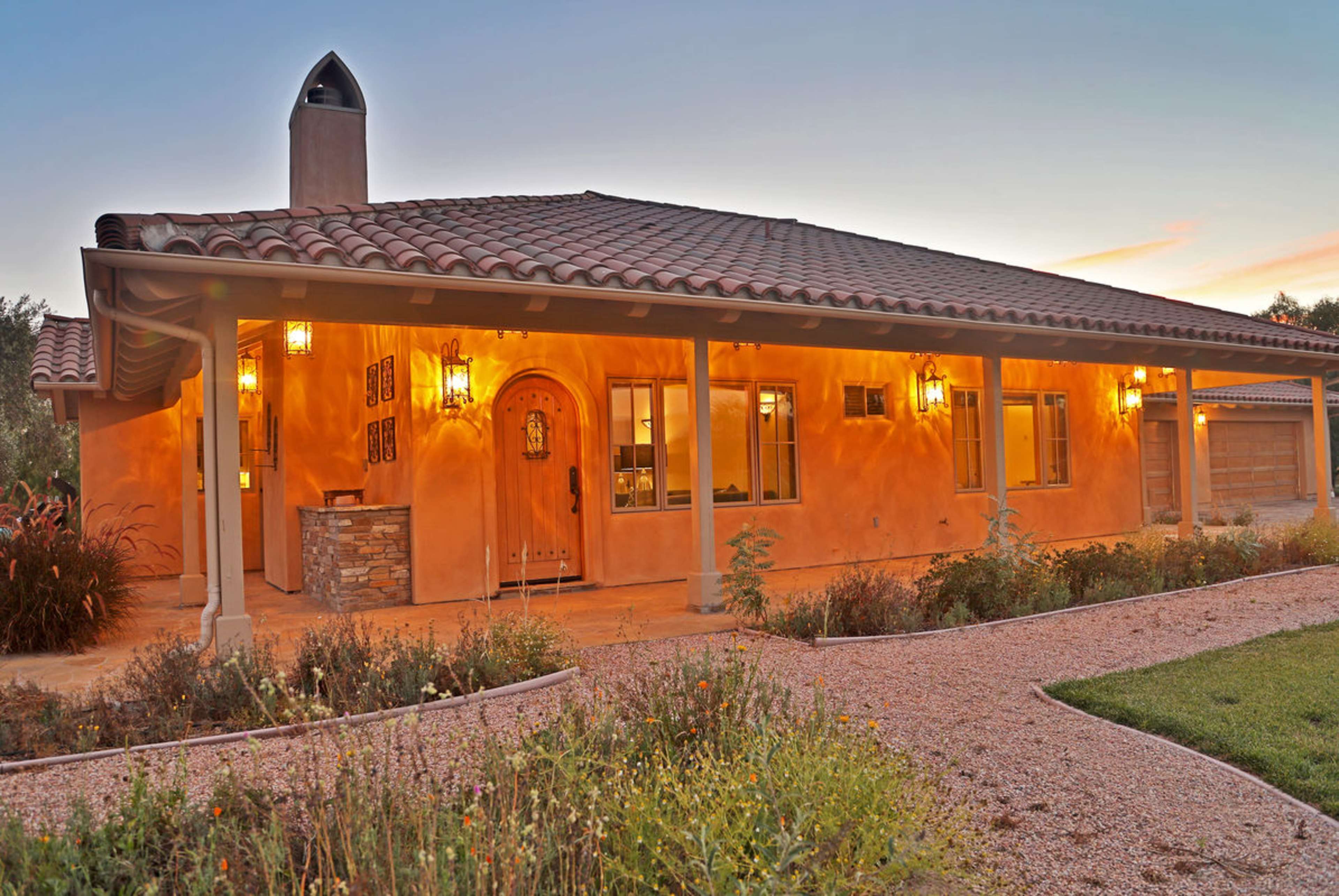 A single-story adobe-style house with a tiled roof and decorative exterior lighting, surrounded by a landscaped yard.