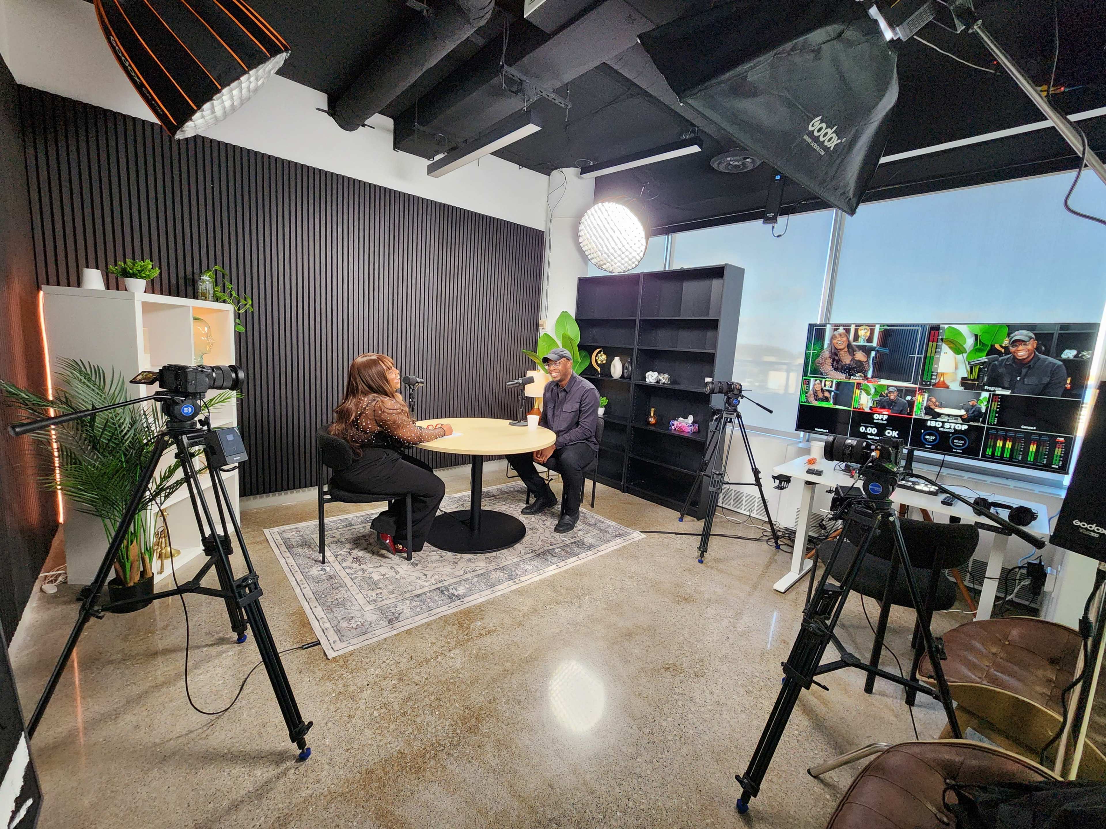 Two individuals are seated at a round table in a modern studio setup, with cameras and production equipment positioned around them.