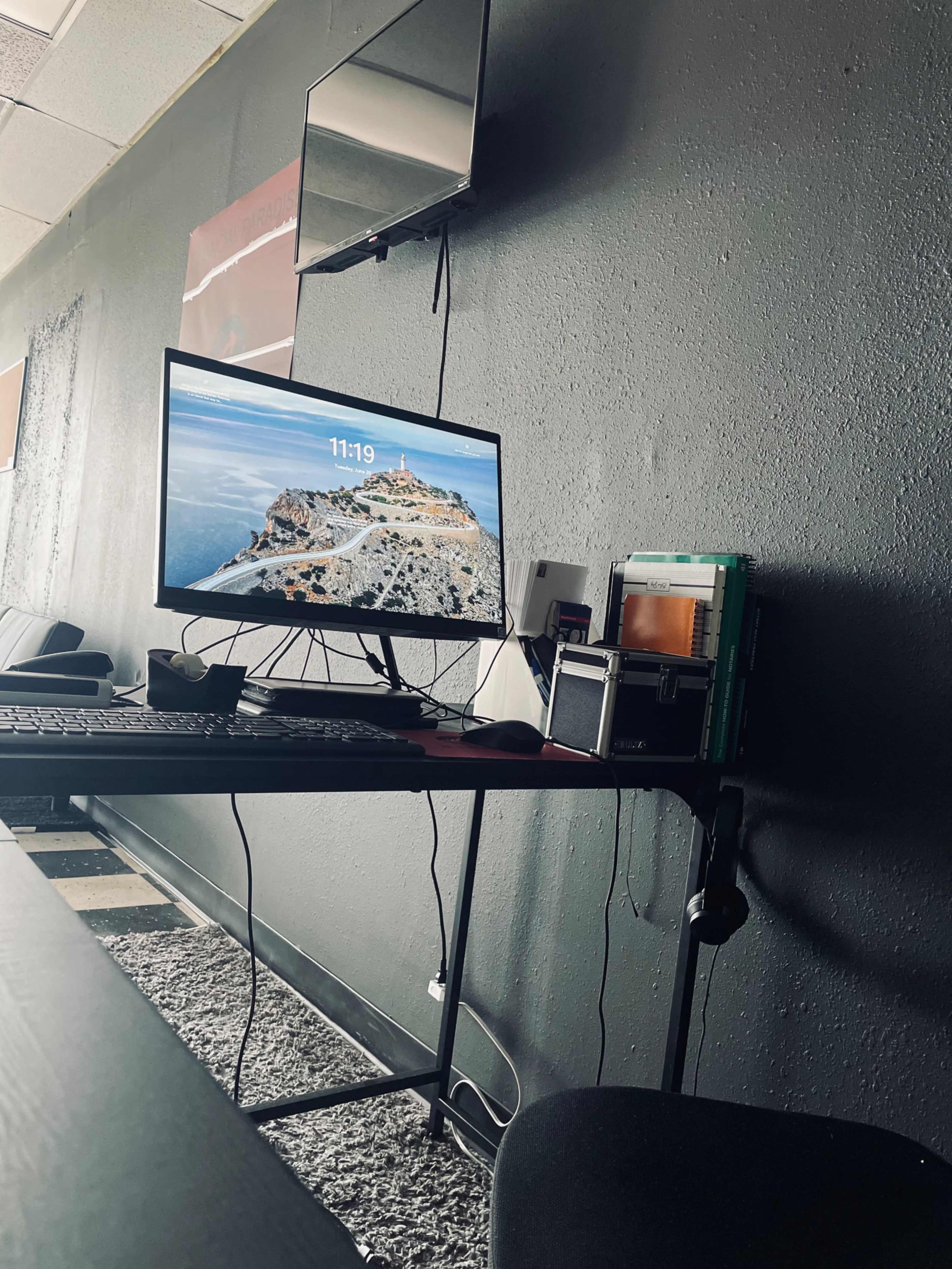 A computer monitor displays the time and a coastal landscape, positioned on a desk alongside a stack of books and a small storage box in an office setting.