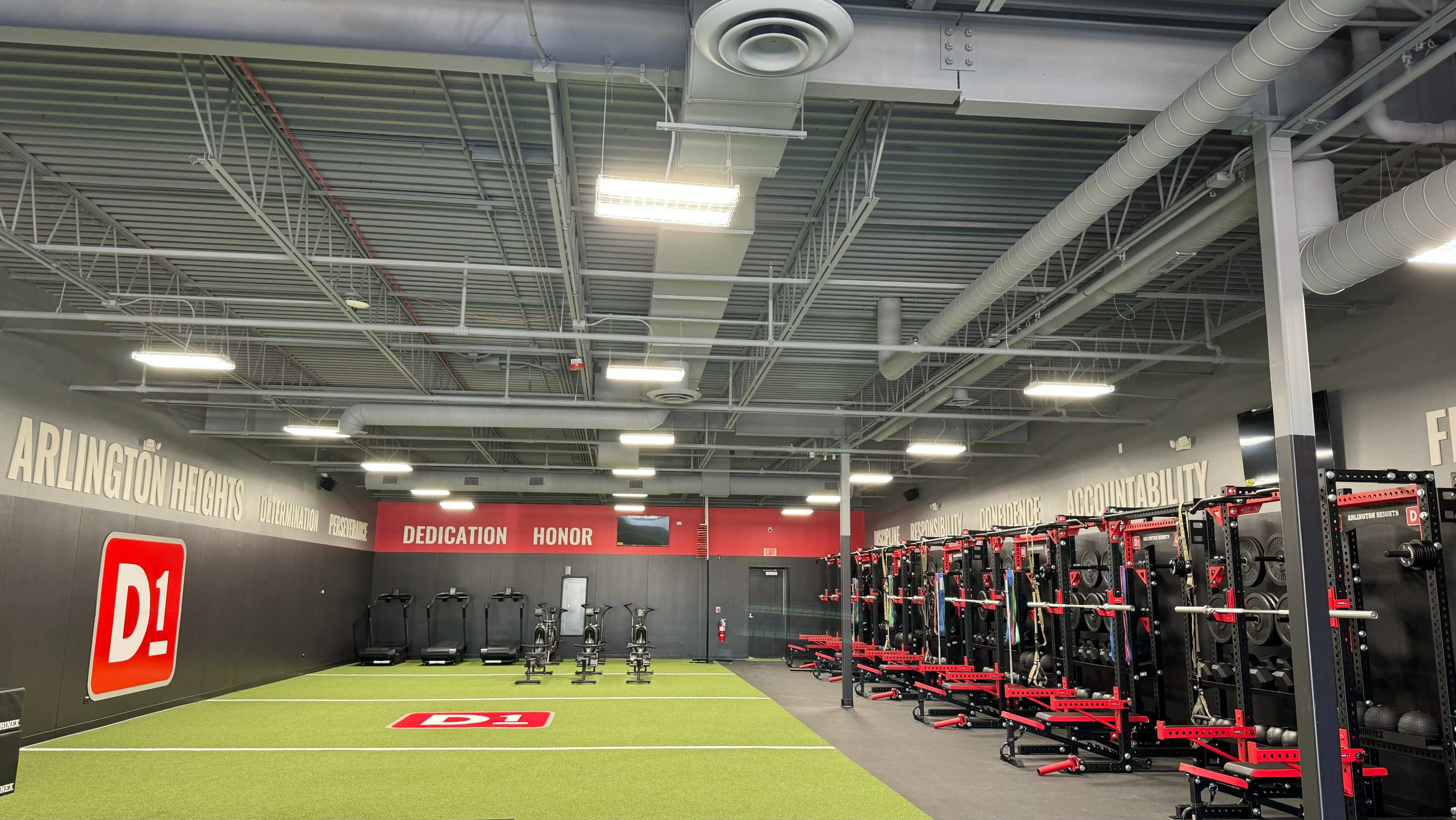 The image shows a modern training facility with workout equipment lined against the walls and green turf flooring.