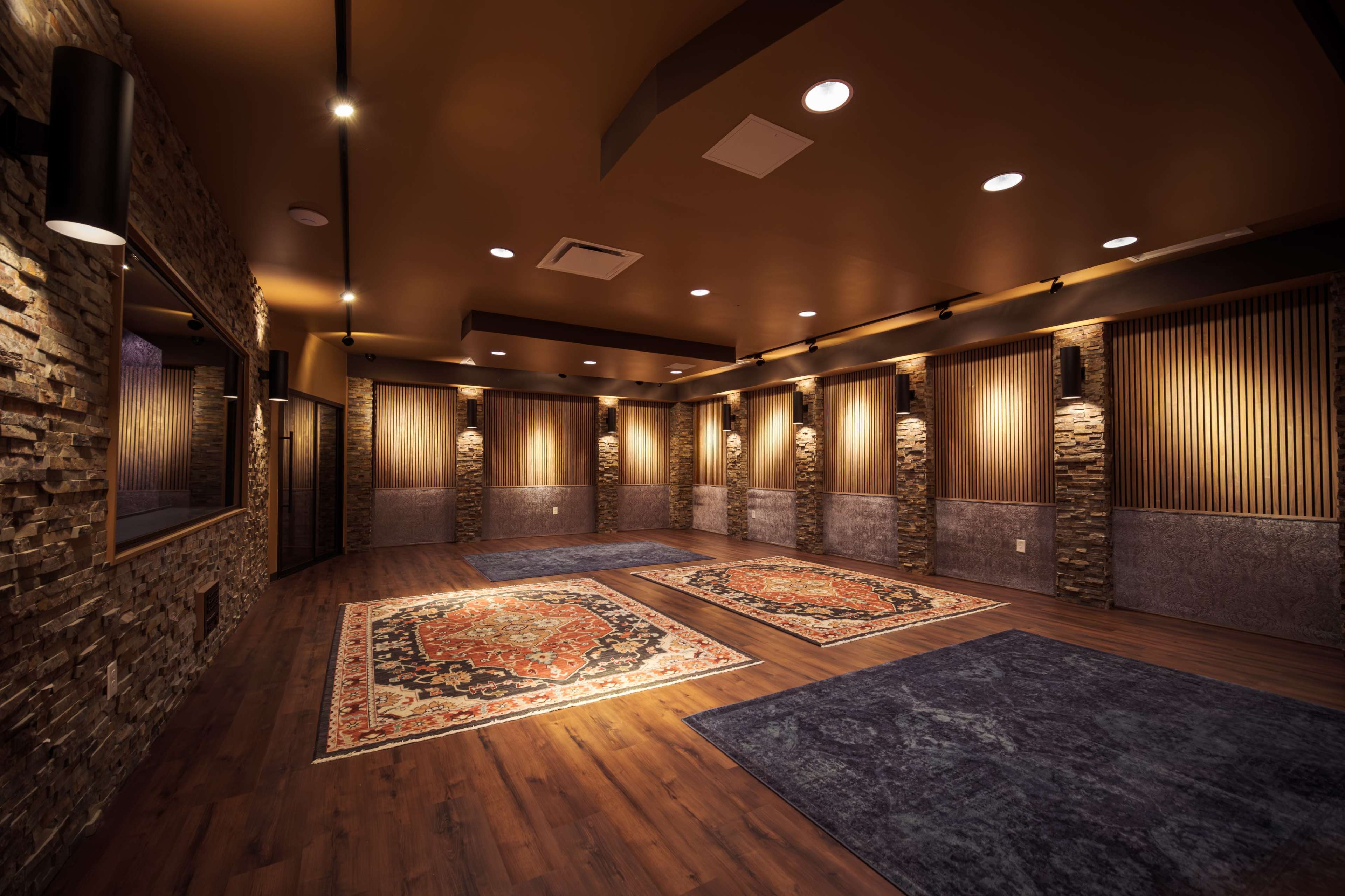 The image shows a spacious, well-lit room with wooden flooring, featuring stone wall segments and several area rugs on the floor.