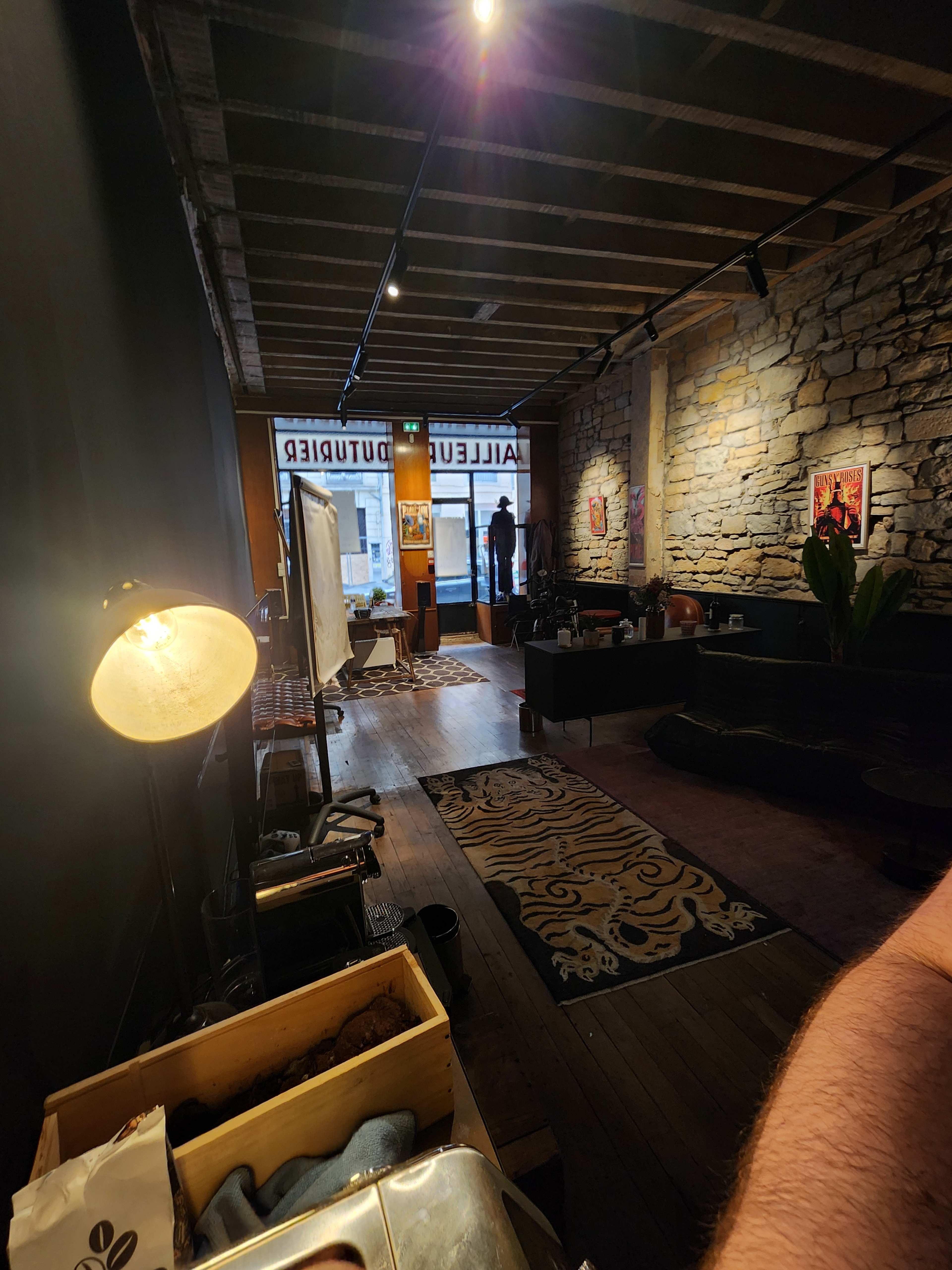 A cozy interior of a café features stone walls, wooden flooring, and a window displaying the street outside, with a person standing near the entrance.