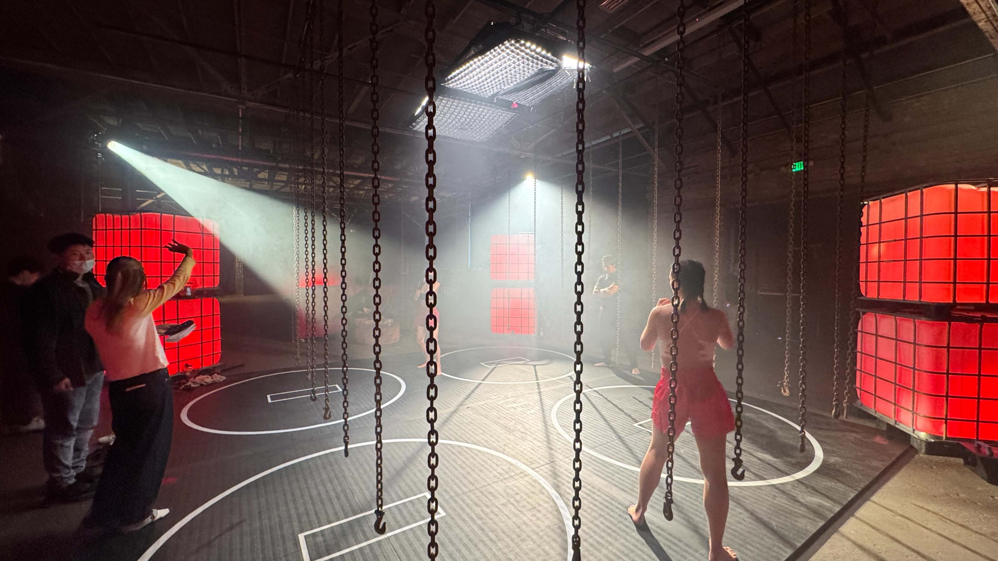 The scene depicts a dimly lit industrial space with chains hanging from the ceiling, where a person in a red outfit stands in the center, surrounded by onlookers and illuminated by beams of light.