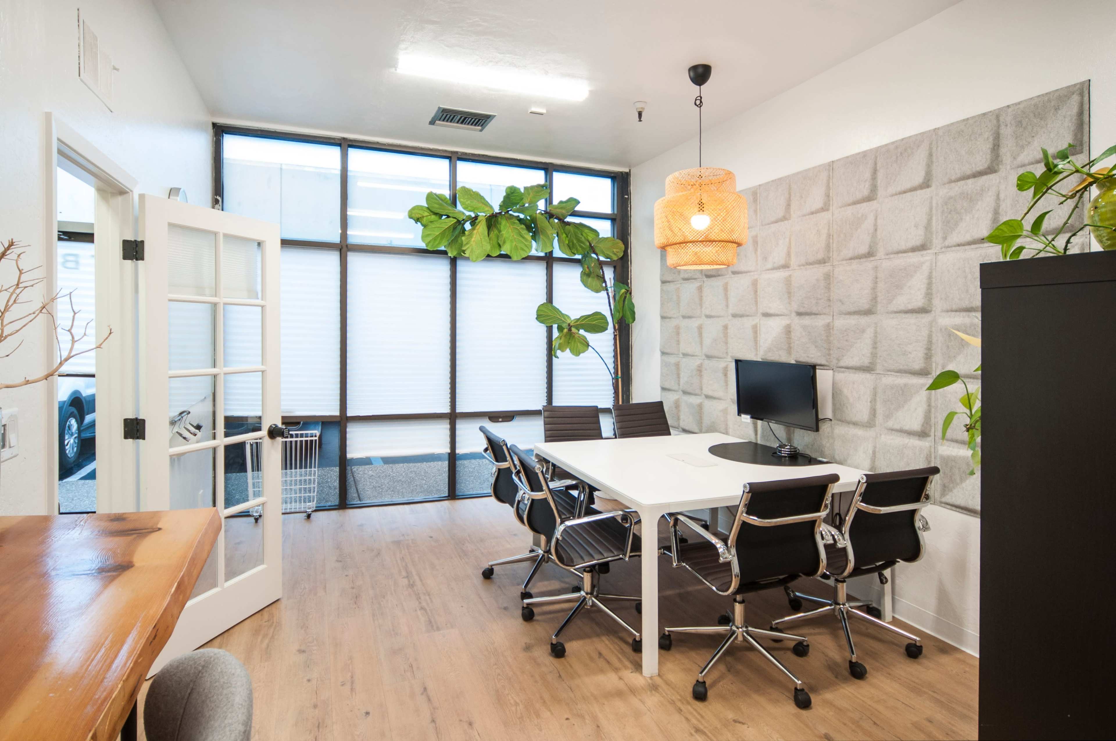 A modern meeting room features a large white table, black chairs, a wall of textured panels, and a window with plants visible outside.