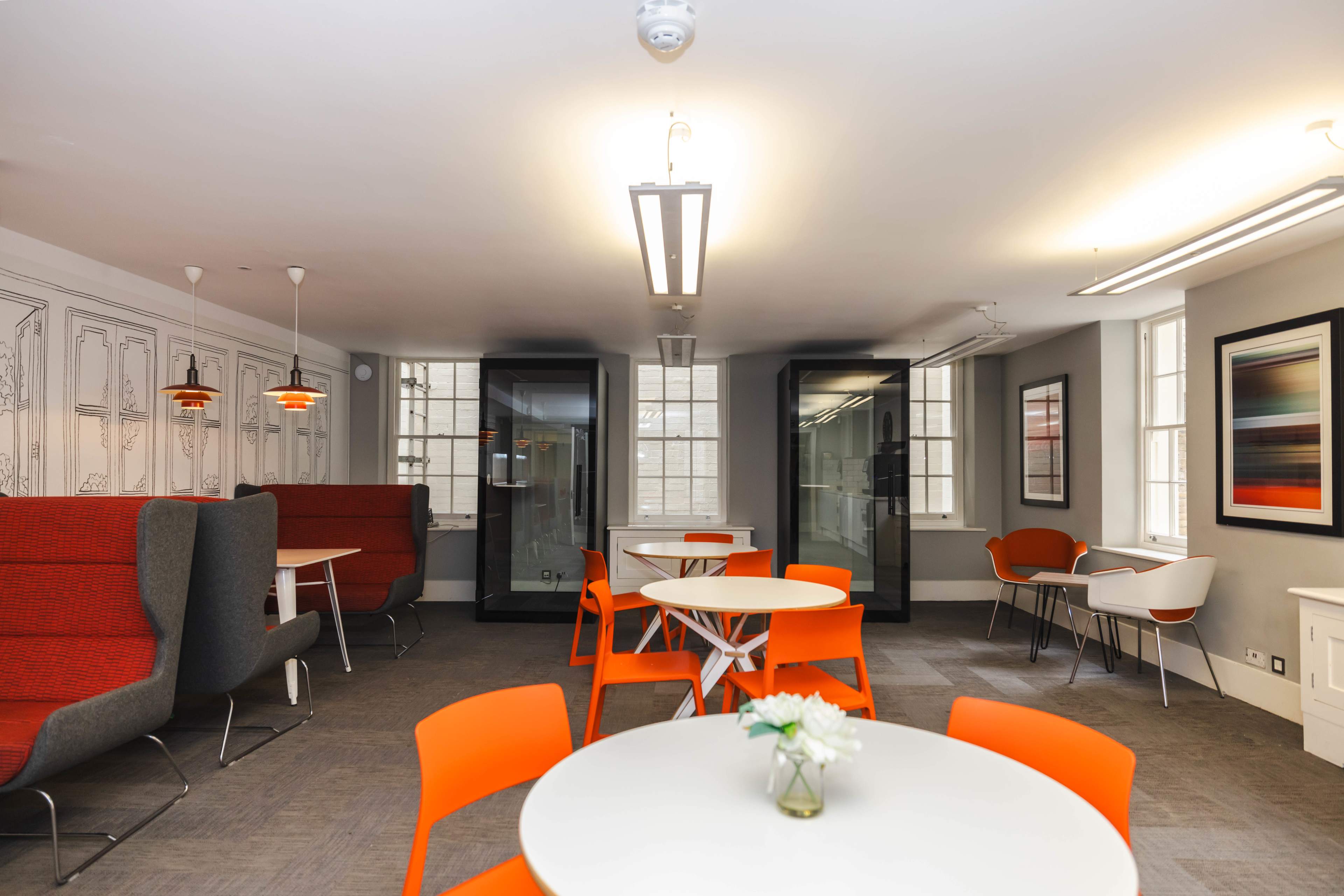 A modern office lounge area with white tables, orange chairs, and soundproof booths, featuring large windows and decorative wall art.