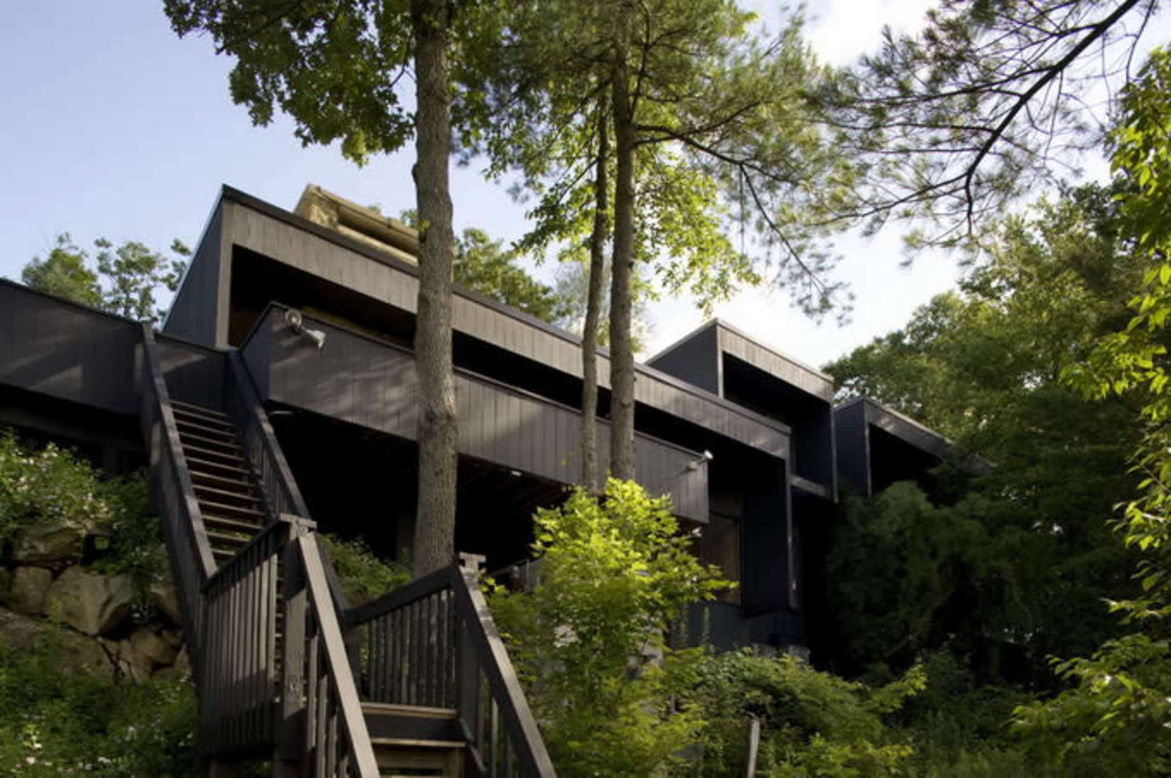 A modern, dark-colored house built on a hillside, surrounded by trees and greenery, with a wooden staircase leading up to it.