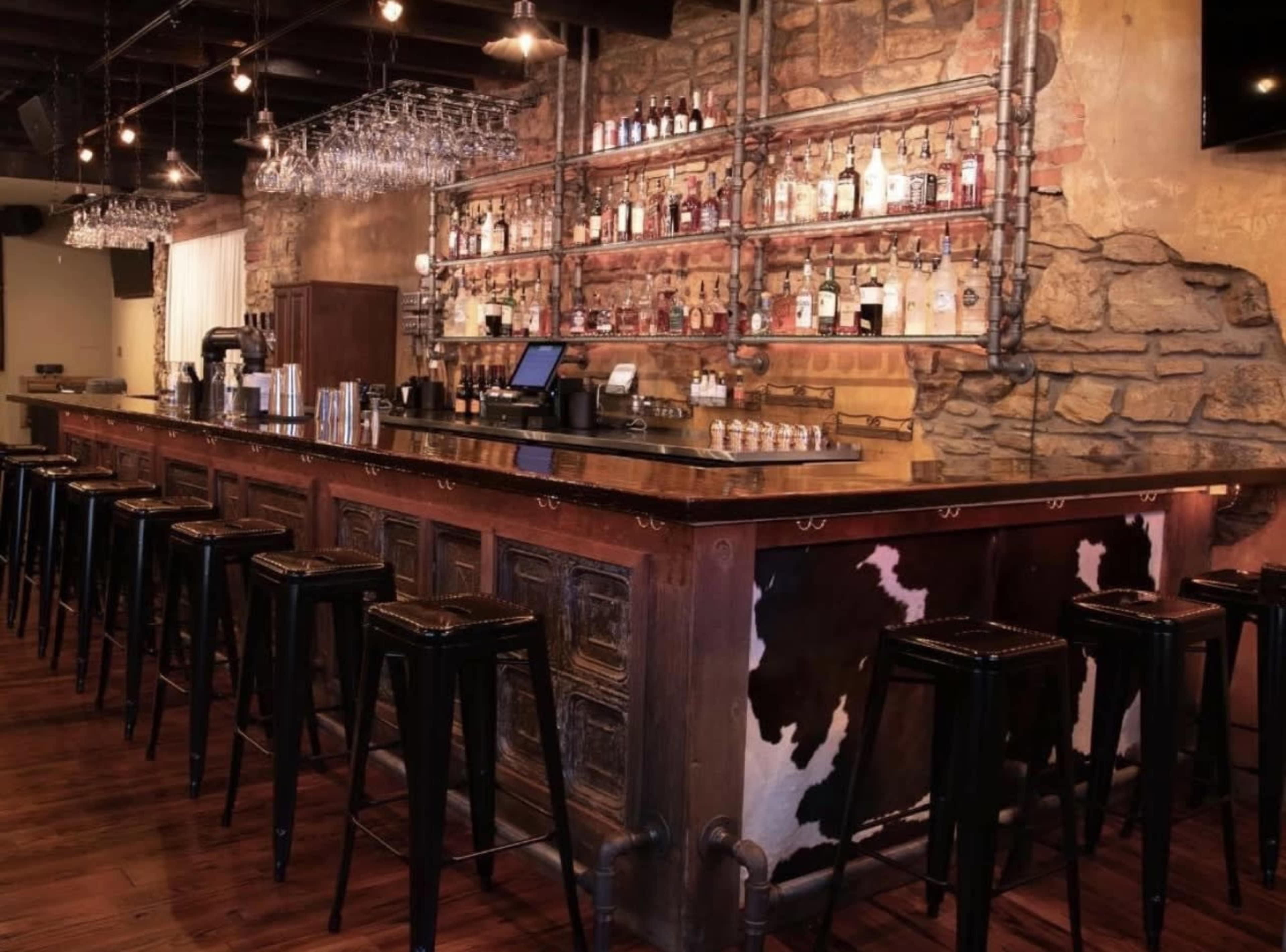A rustic bar with a polished countertop, wooden stools, and shelves displaying a variety of liquor bottles against a stone wall.