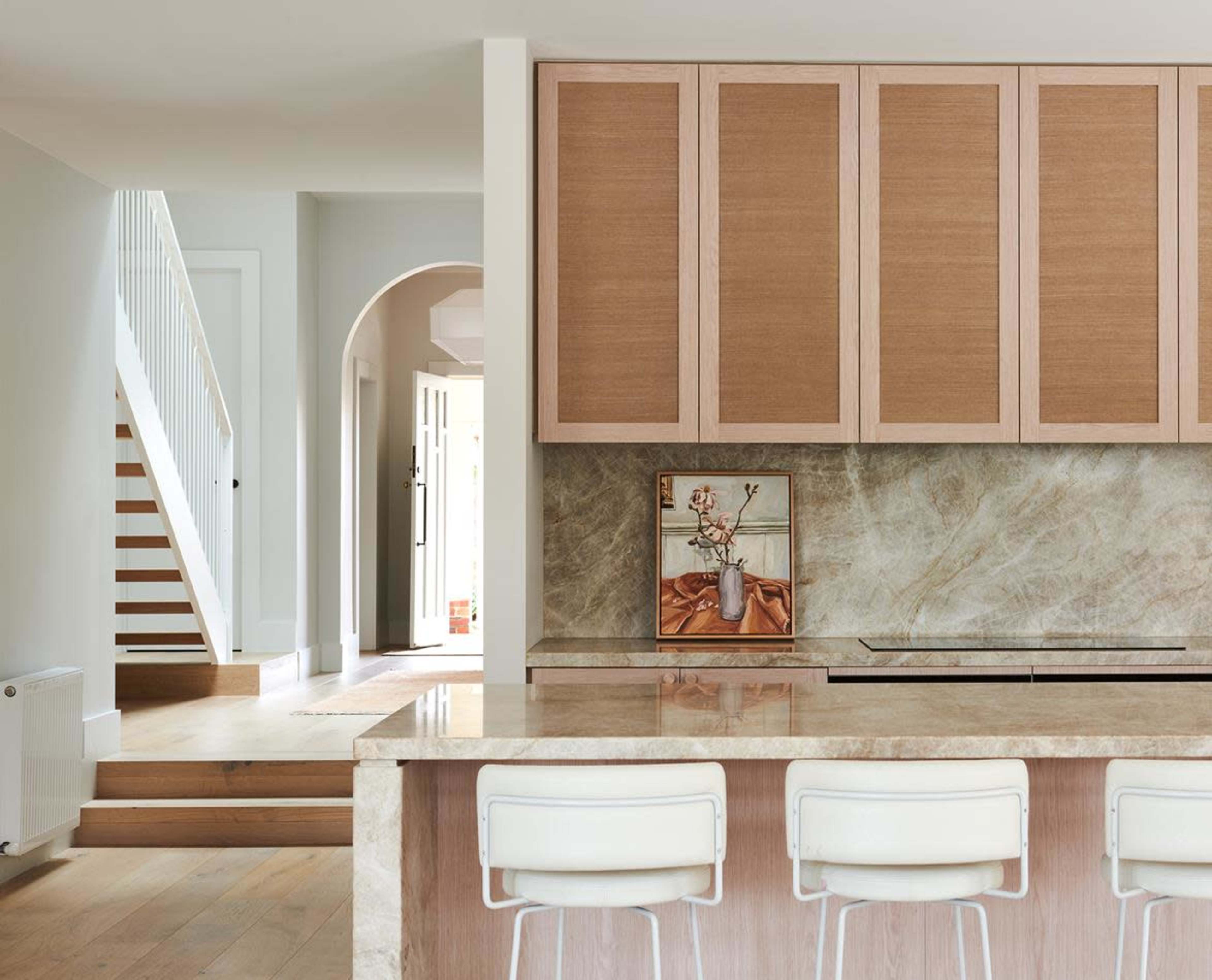 A modern kitchen with light wood cabinetry, a marble countertop, and a staircase leading to another area of the home.