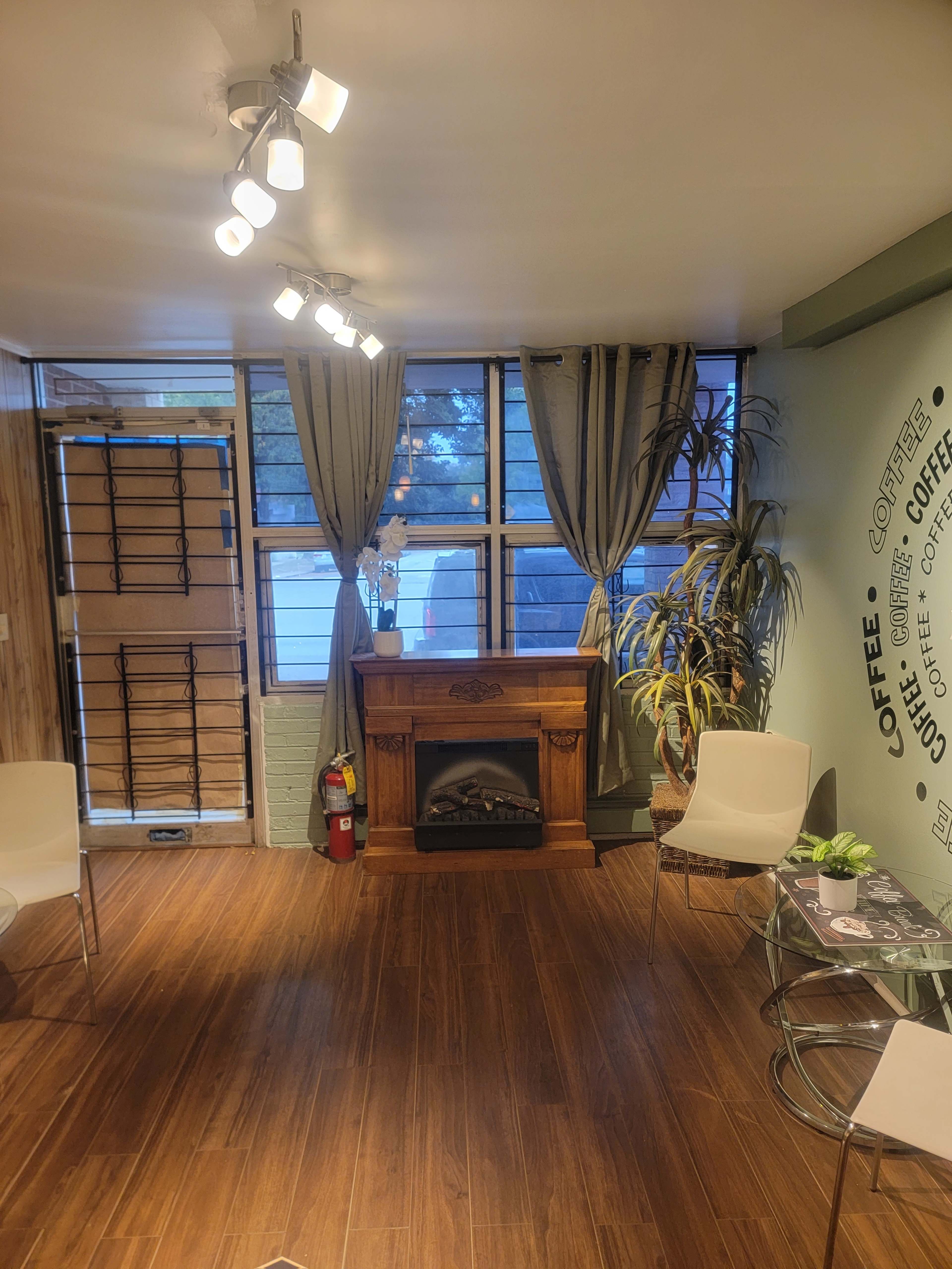 A cozy room featuring a decorative fireplace, large windows with curtains, wooden flooring, and two chairs surrounding a glass table.