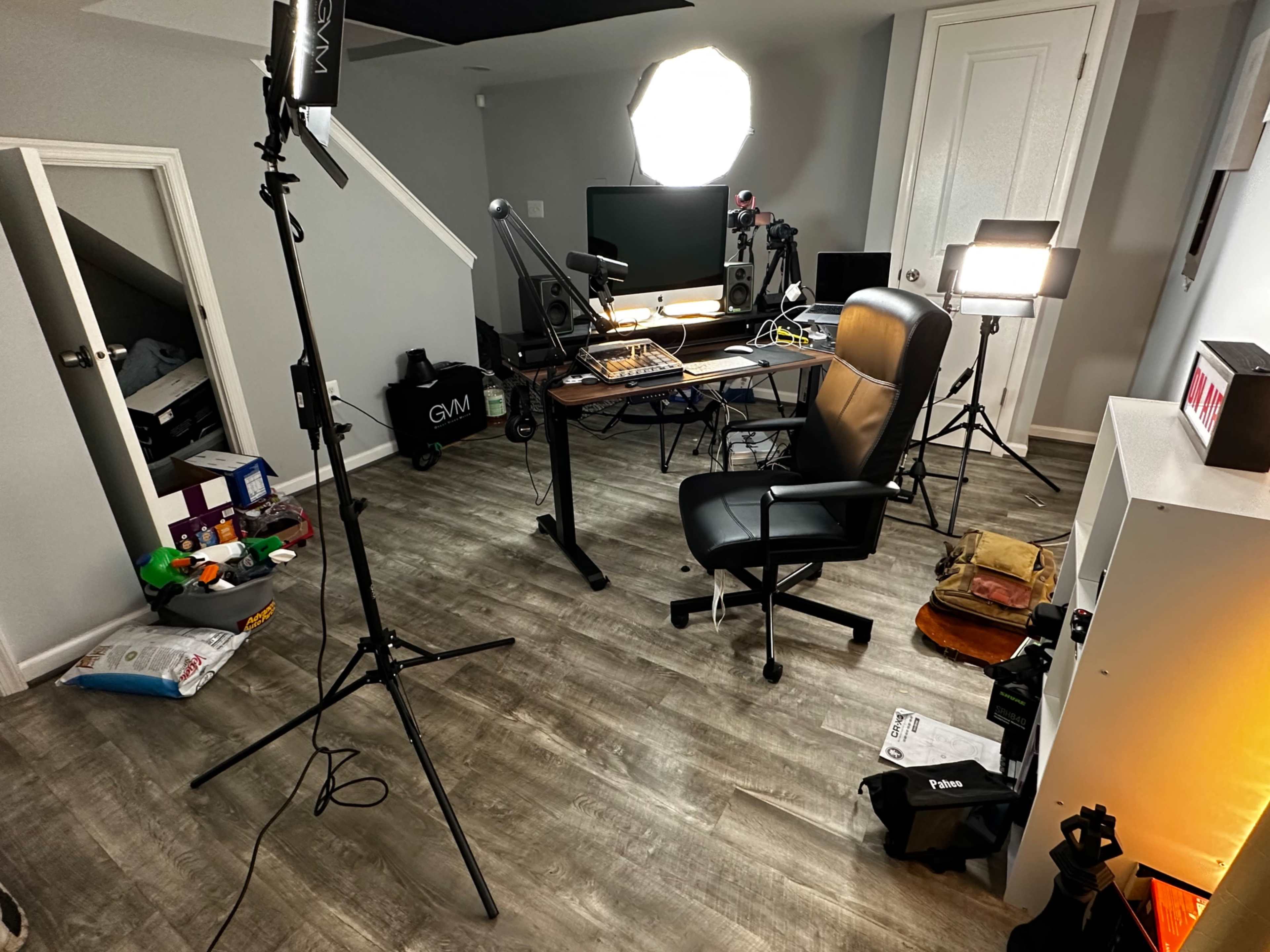 A cluttered home office setup features a desk with computer equipment, lighting stands, and scattered items on the floor.