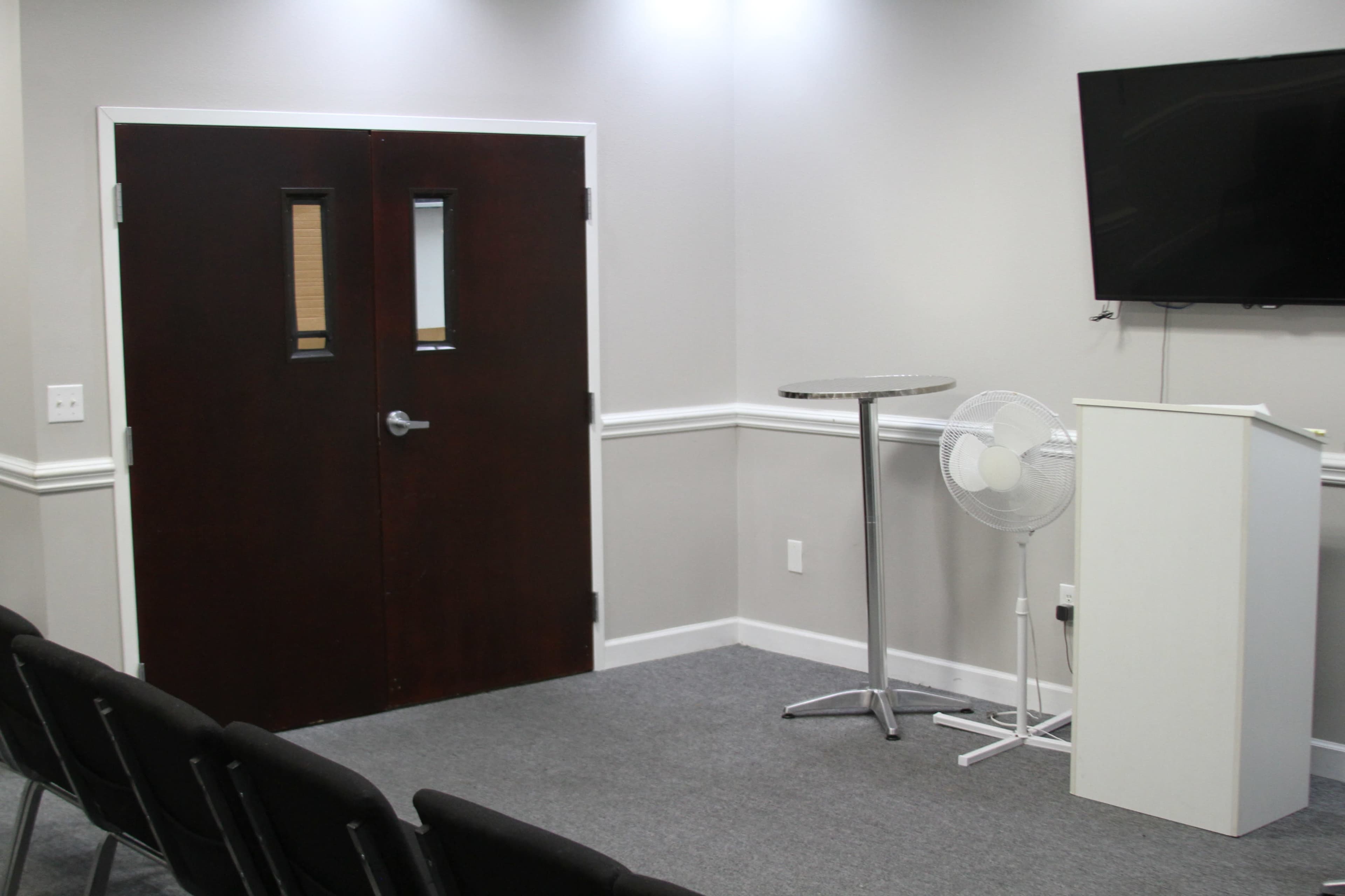 The image depicts a small meeting room featuring a dark wooden door, a podium, a standing fan, and a television mounted on the wall, with rows of chairs arranged in front.