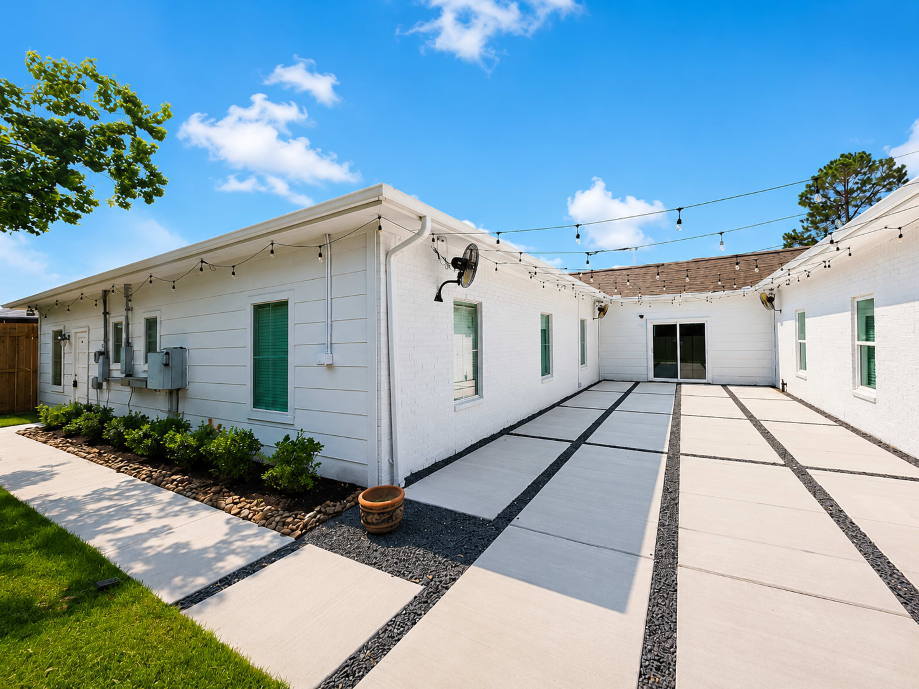 The image shows a landscaped outdoor area of a white residential building with a concrete pathway and string lights overhead.
