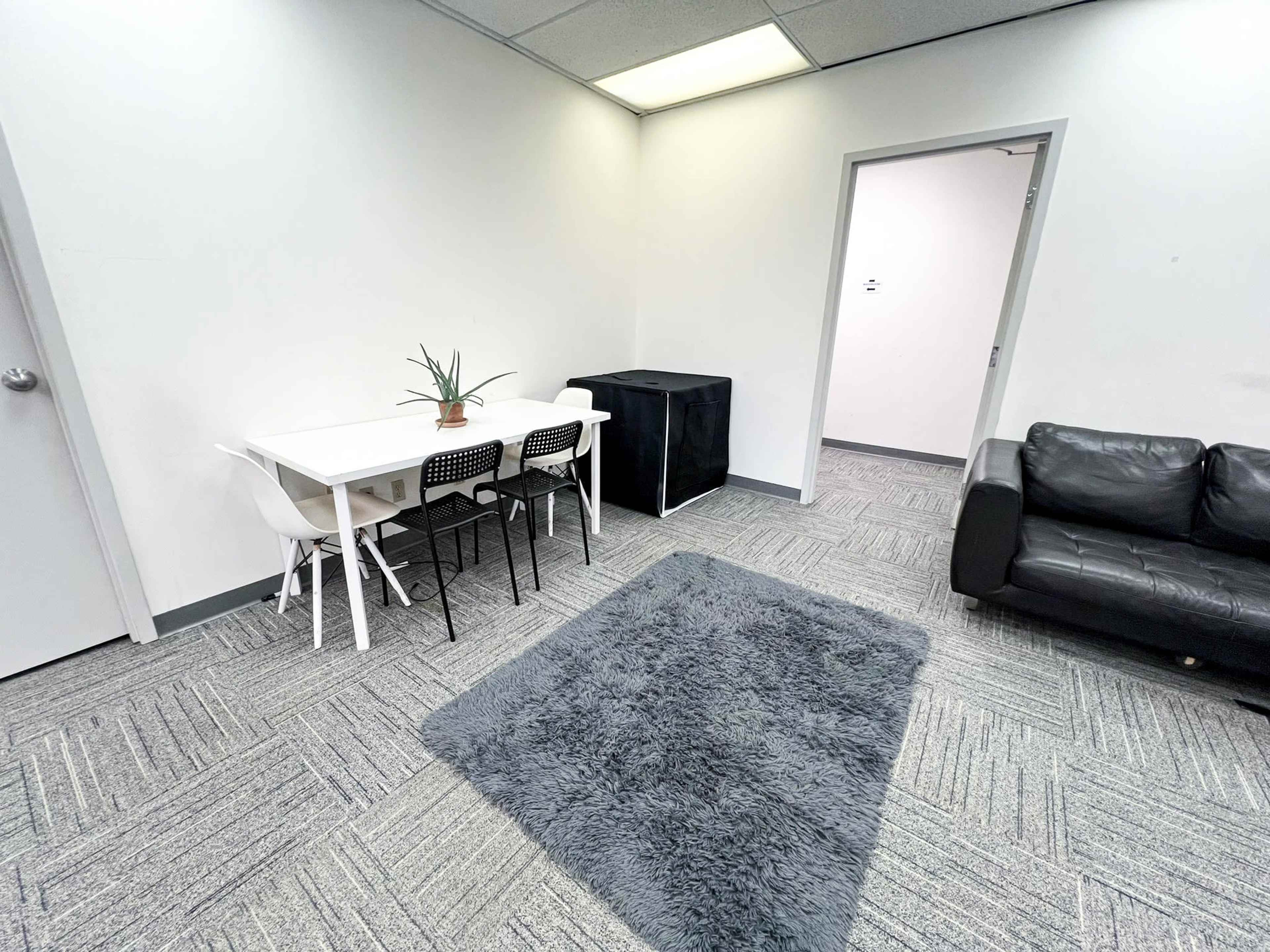 A minimalist office space featuring a white table with four chairs, a black couch, and a gray rug on the carpeted floor.