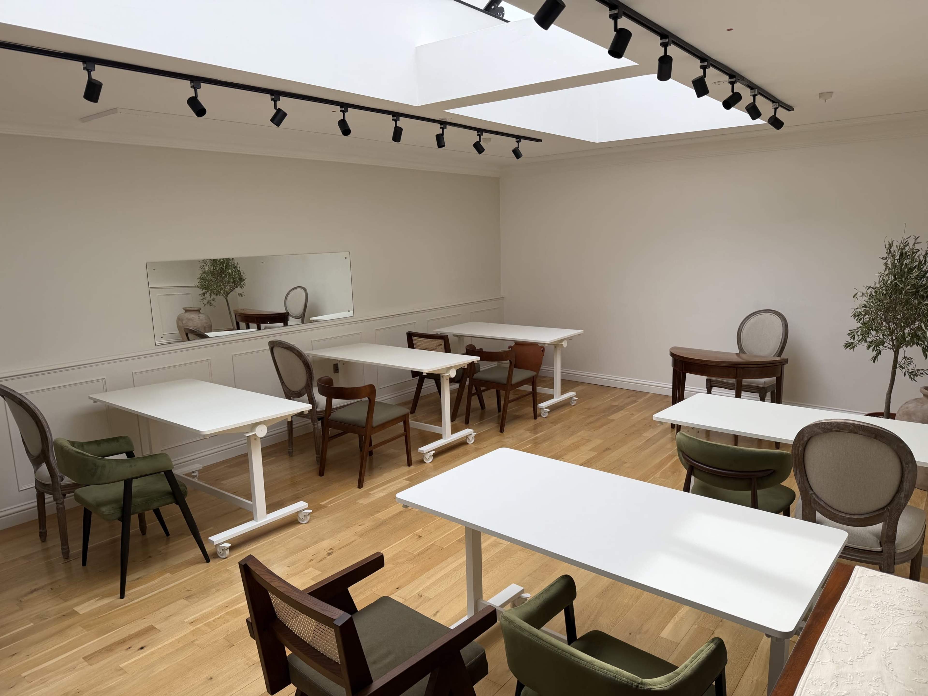 The image shows a spacious room with wooden floors featuring several tables and chairs arranged in an organized manner, along with a large mirror on one wall and a skylight above.