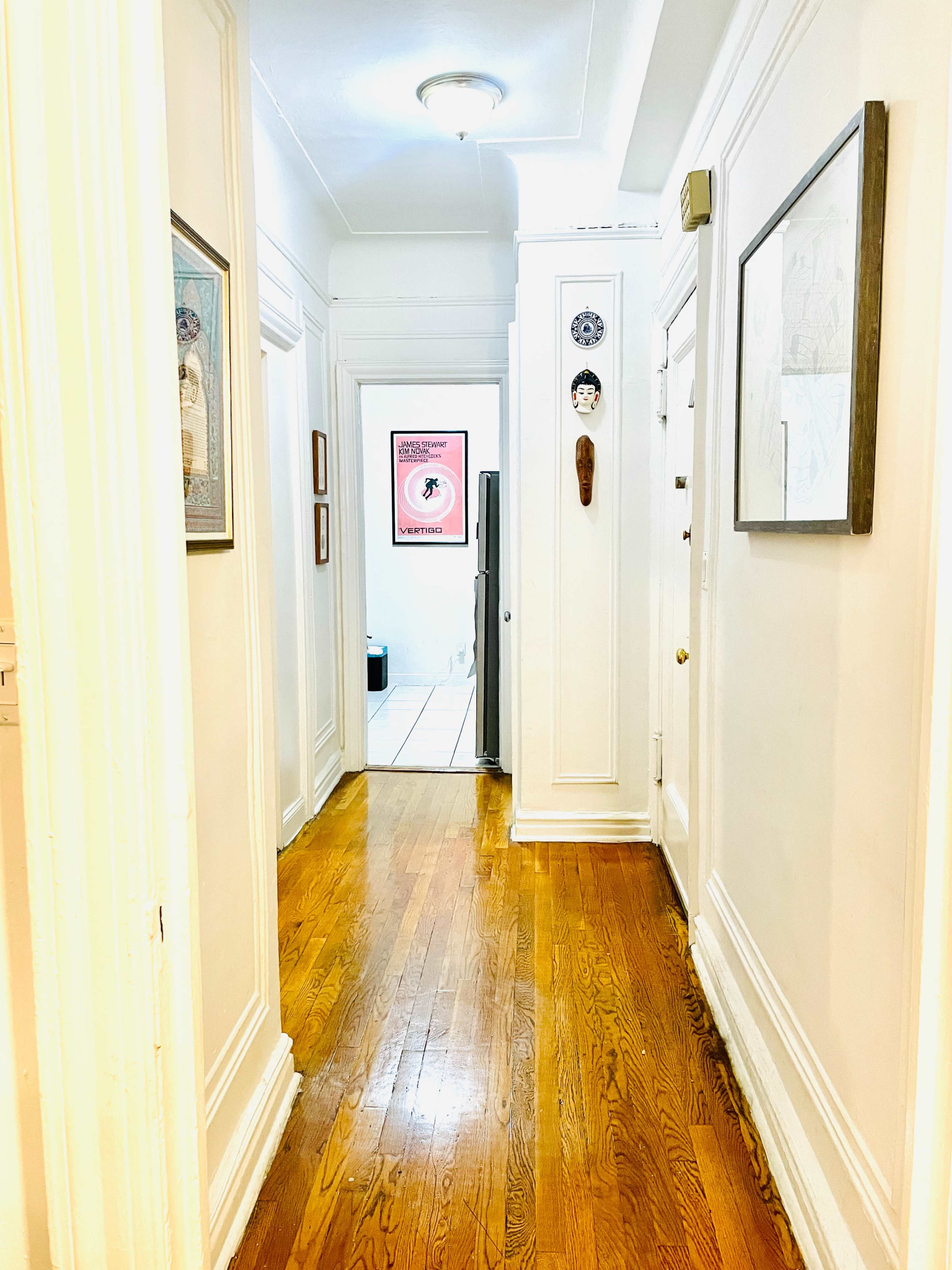 A narrow hallway with hardwood flooring leads to a doorway at the end, framed by white walls and decorated with various framed artworks.