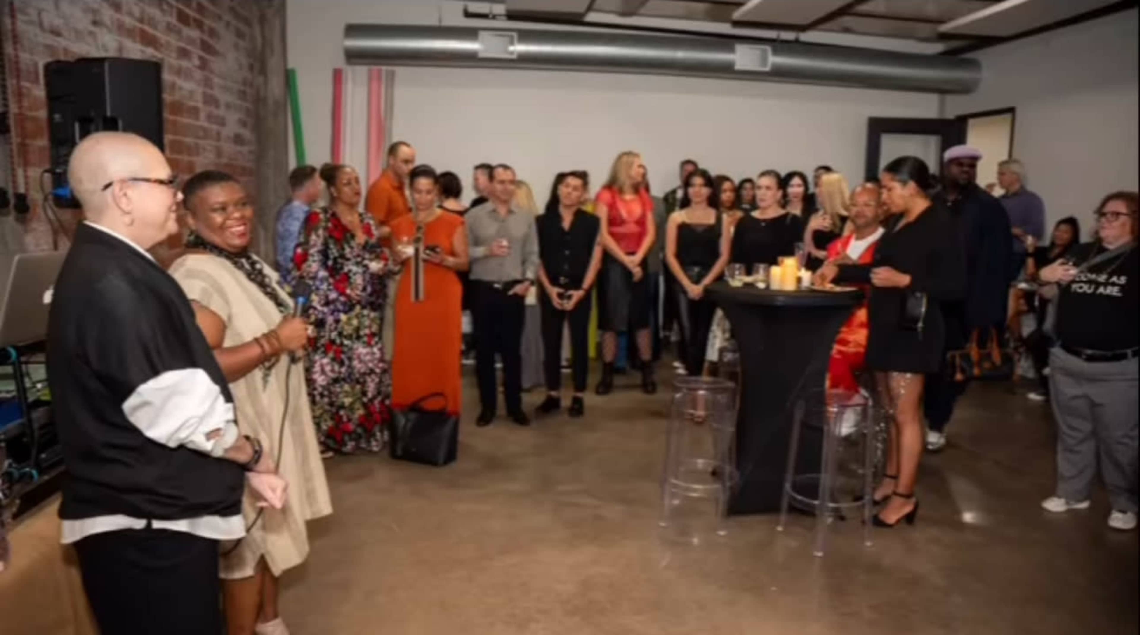 The image shows a lively gathering of people at an indoor event, with attendees standing and socializing around tables in a modern space.