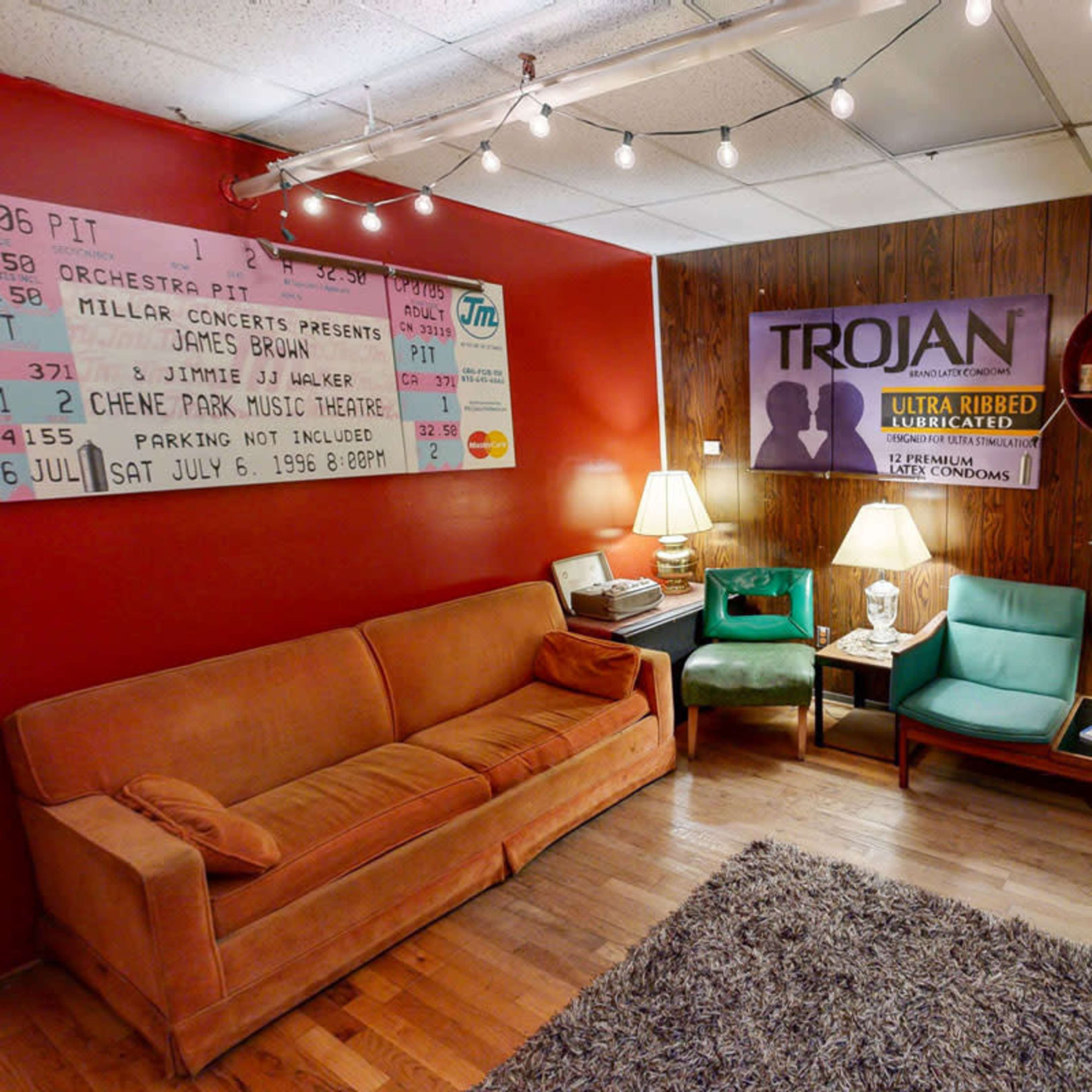 The image shows a small room with a brown couch and two green chairs, decorated with a poster about a concert and a banner for Trojan condoms, with lights hanging overhead.