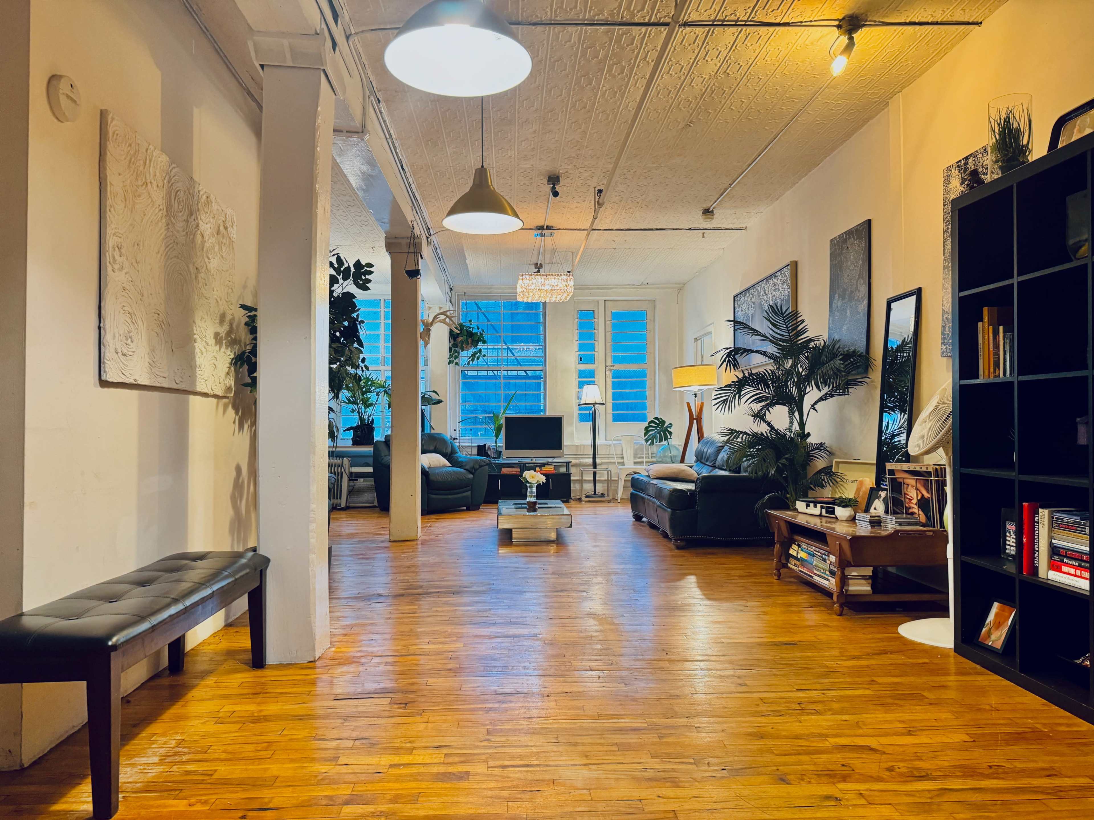 The image shows a spacious, well-lit living area with wooden floors, plants, and various seating arrangements around a coffee table.