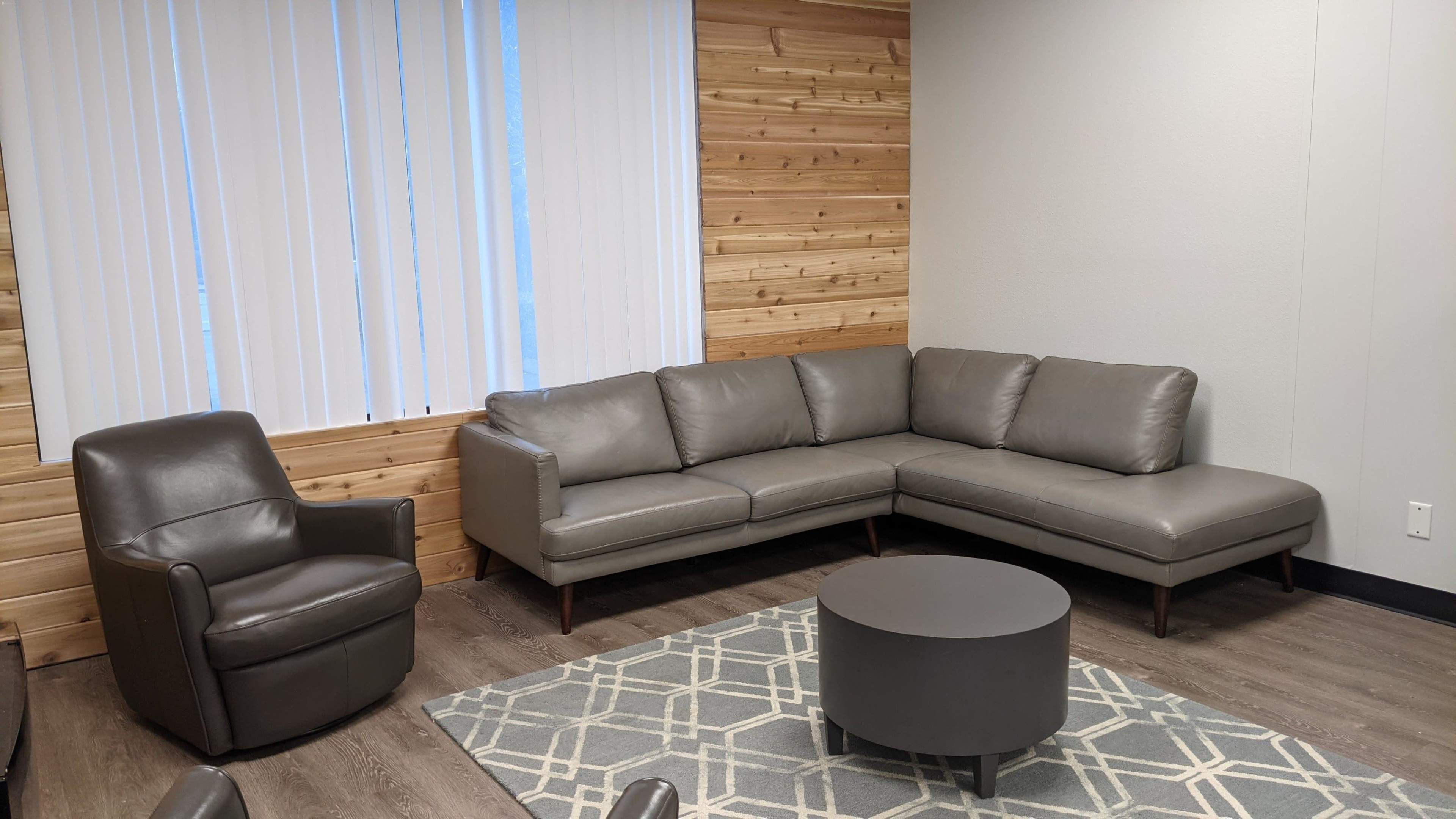 The image shows a modern, cozy living room with a gray sectional sofa, a circular table, and a single armchair, all set against a backdrop of wooden wall panels and a window with vertical blinds.
