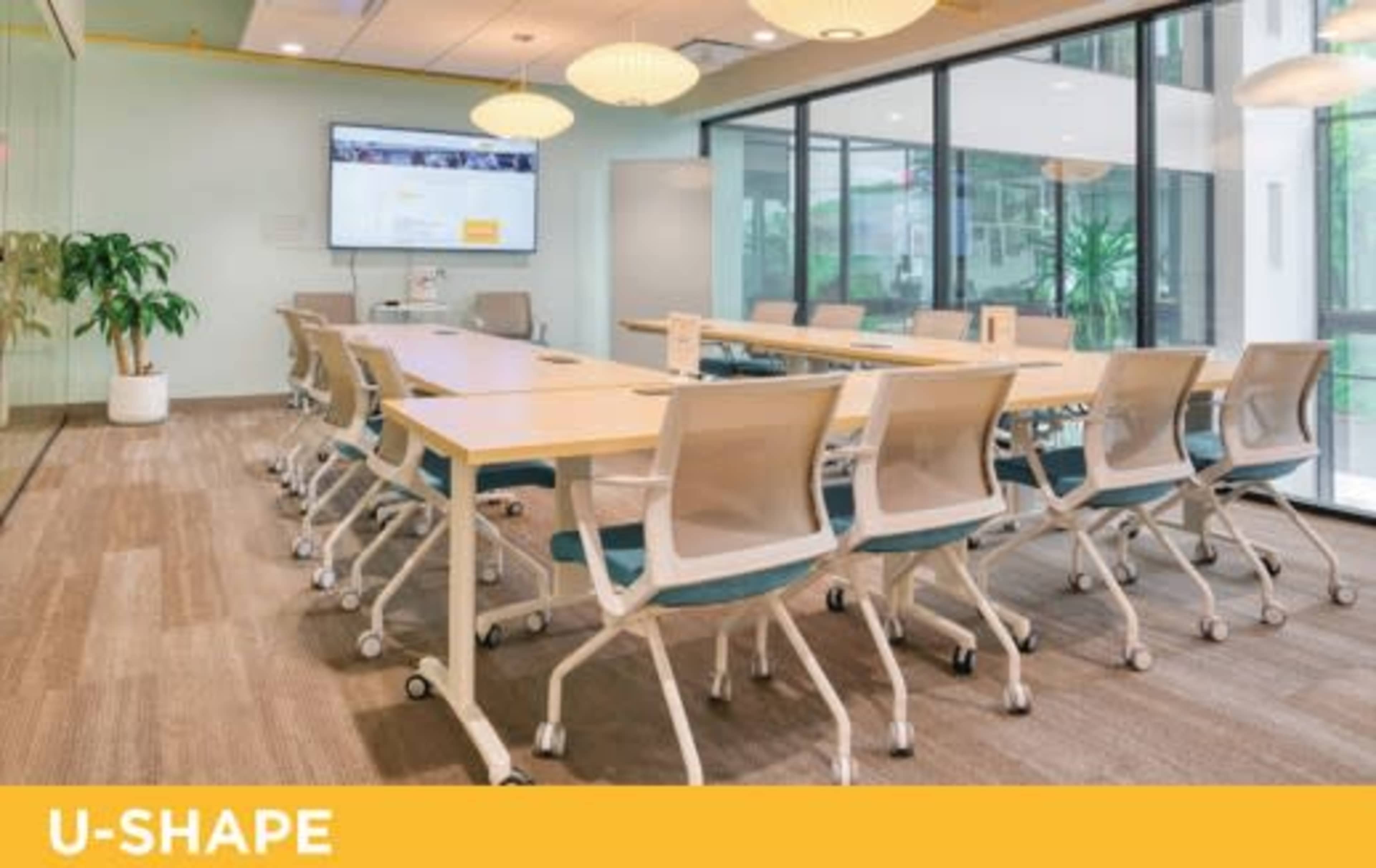 A conference room features a U-shaped arrangement of tables and chairs, with large windows providing natural light and a screen displaying content at the front.