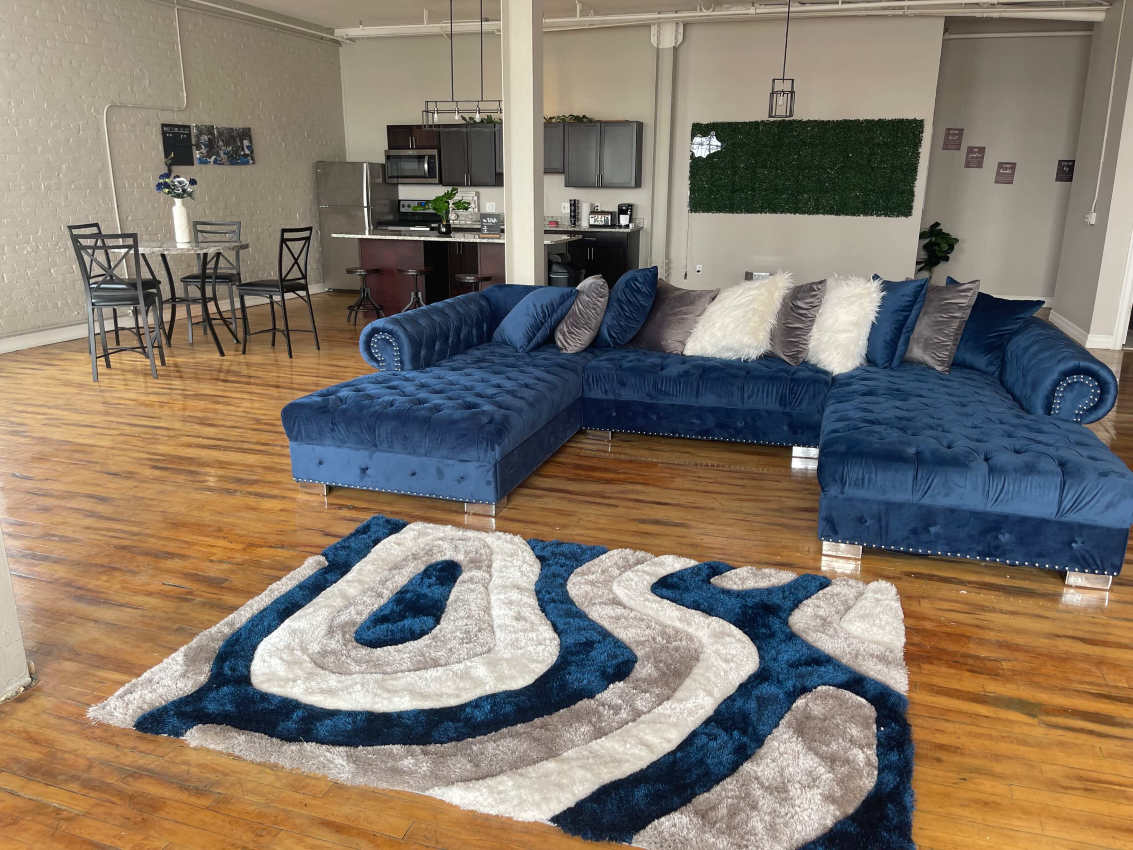The image shows a spacious living area featuring a blue sectional sofa with decorative cushions, a patterned rug, a small dining table, and a modern kitchen in the background.