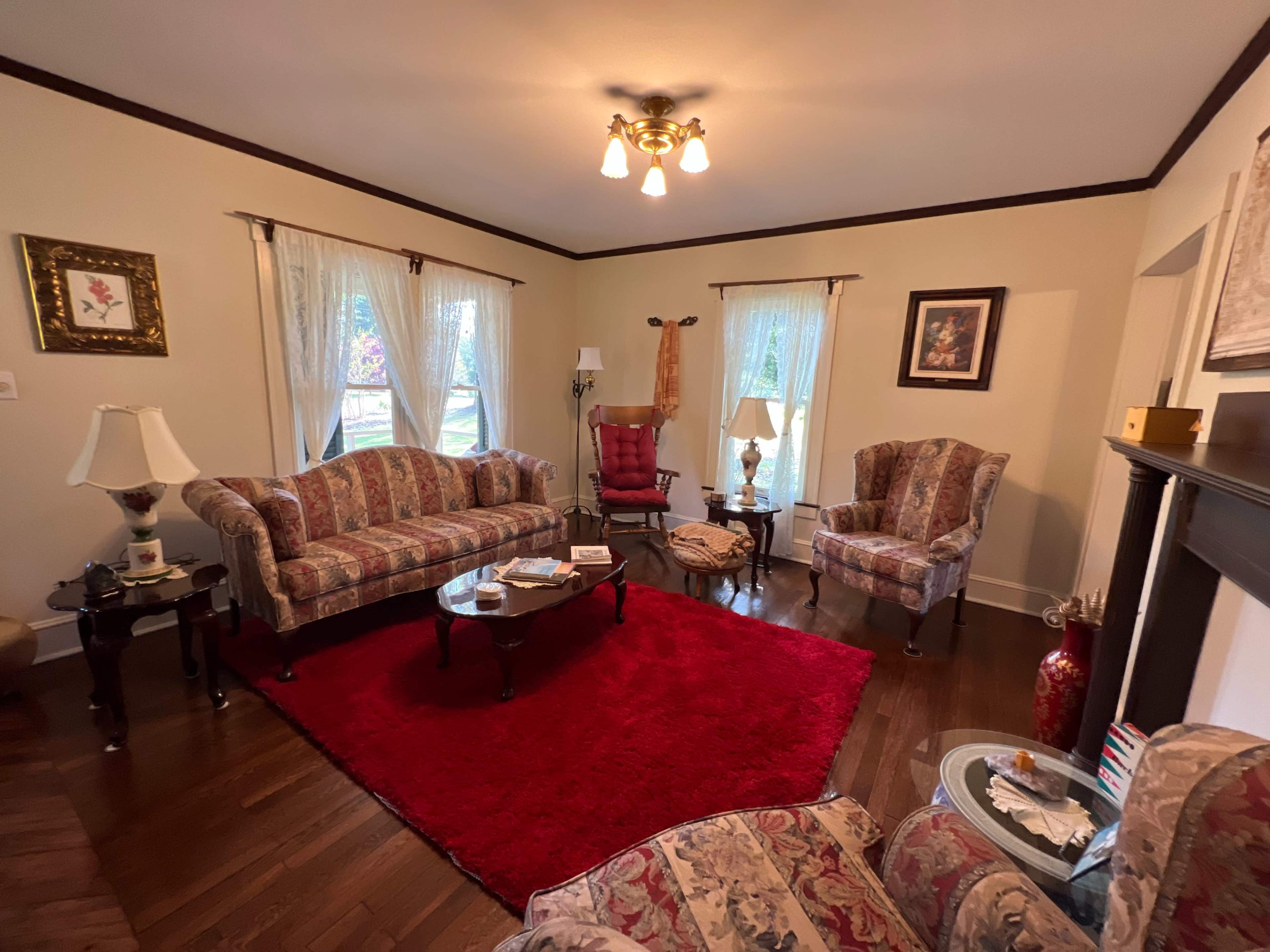 The living room features patterned furniture, a red area rug, and large windows allowing natural light to enter.