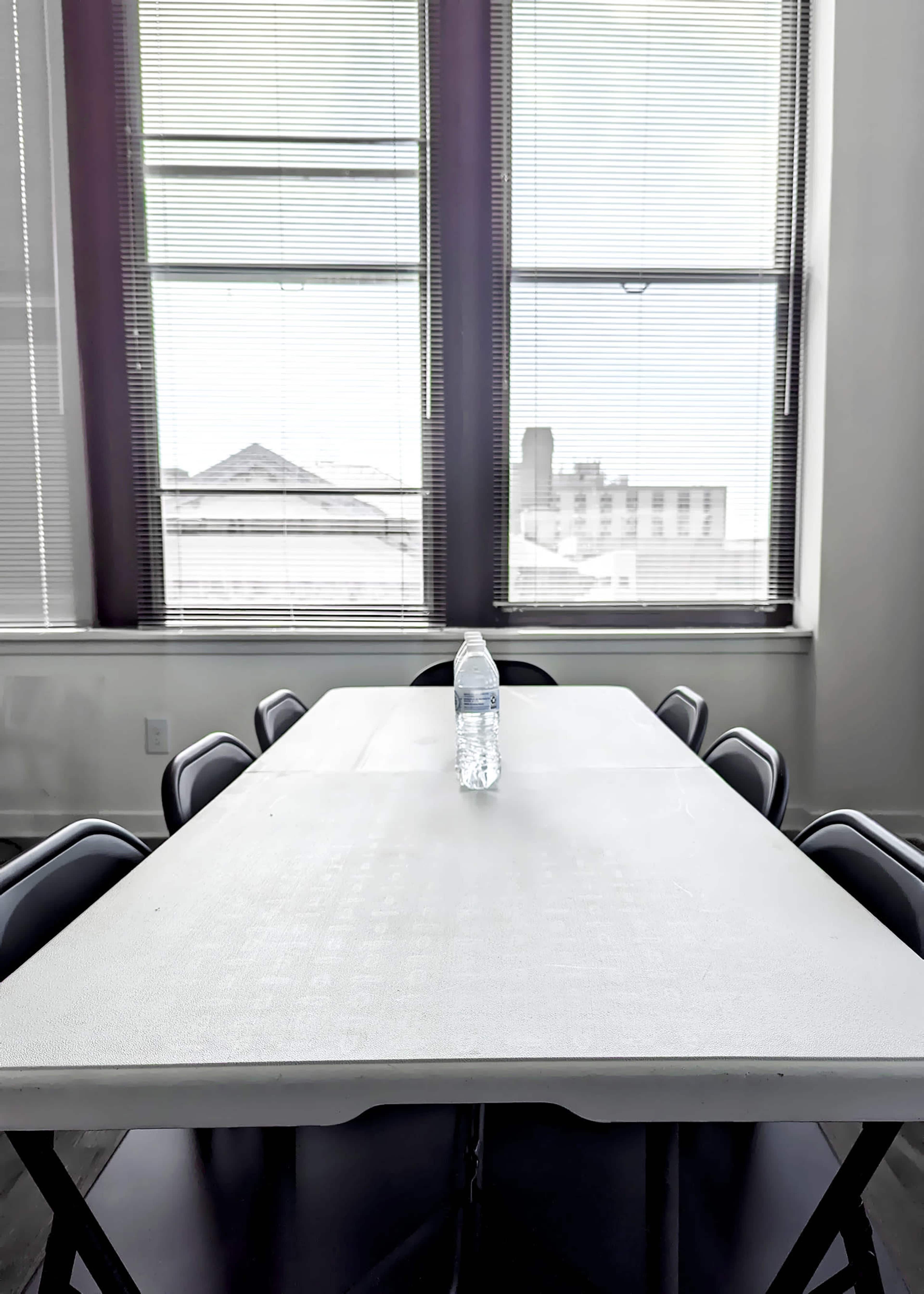 A long, white table is set in a room with large windows, featuring a single water bottle placed in the center.