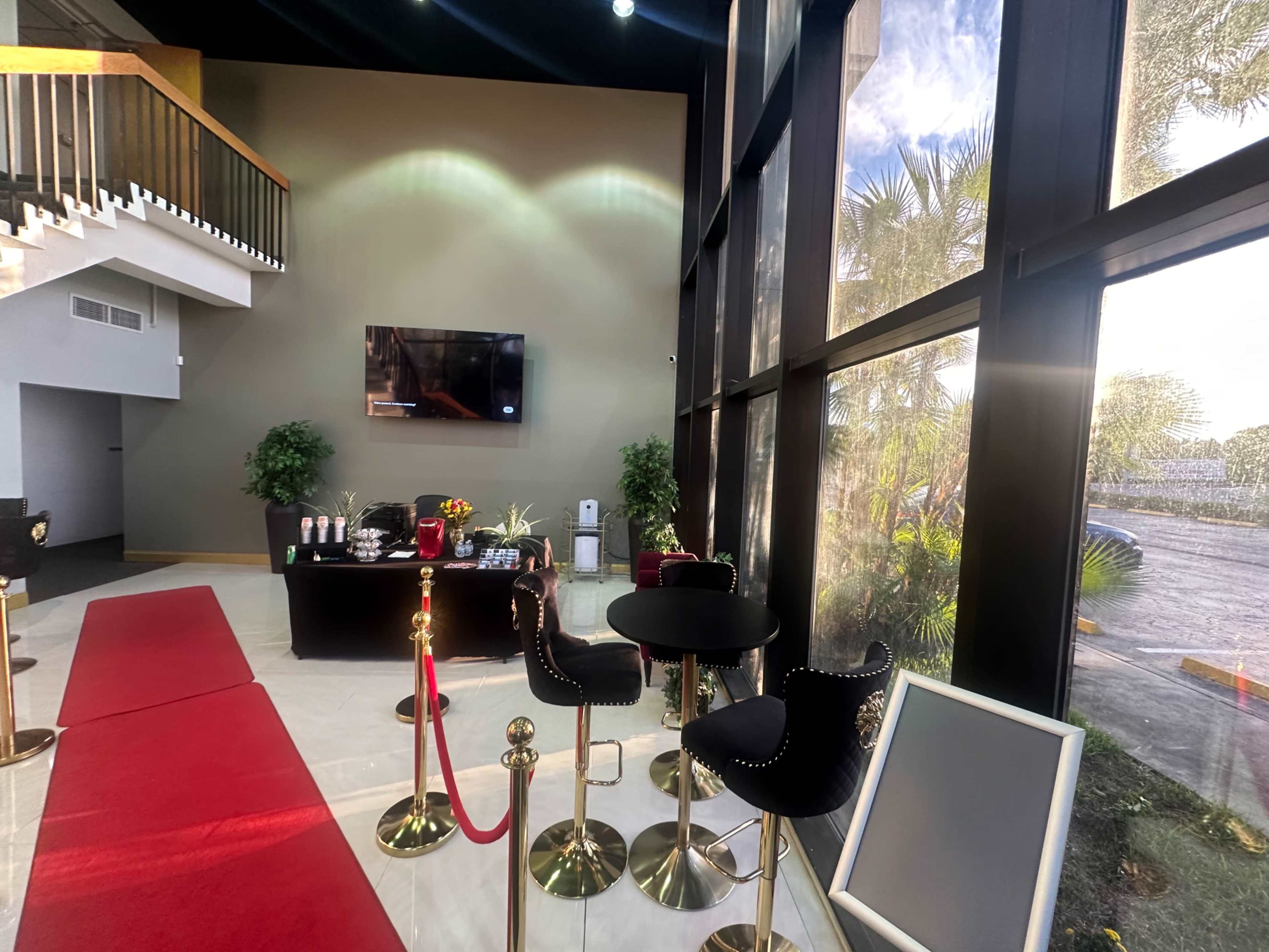 The image shows a modern lobby area with a red carpet, a seating arrangement with black chairs and tables, and large windows letting in natural light.