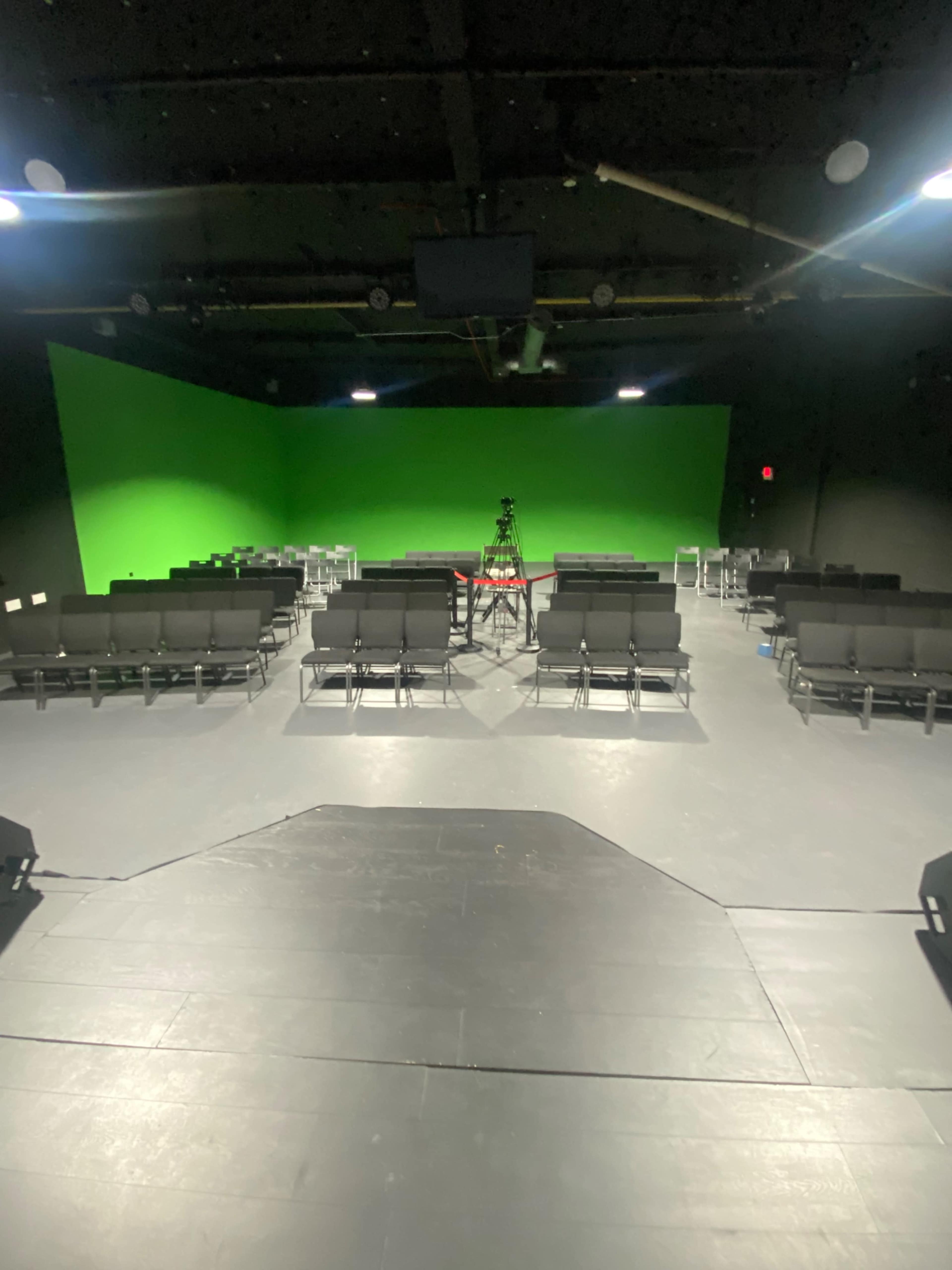 A theater space features rows of black chairs facing a center stage, with a green screen backdrop.