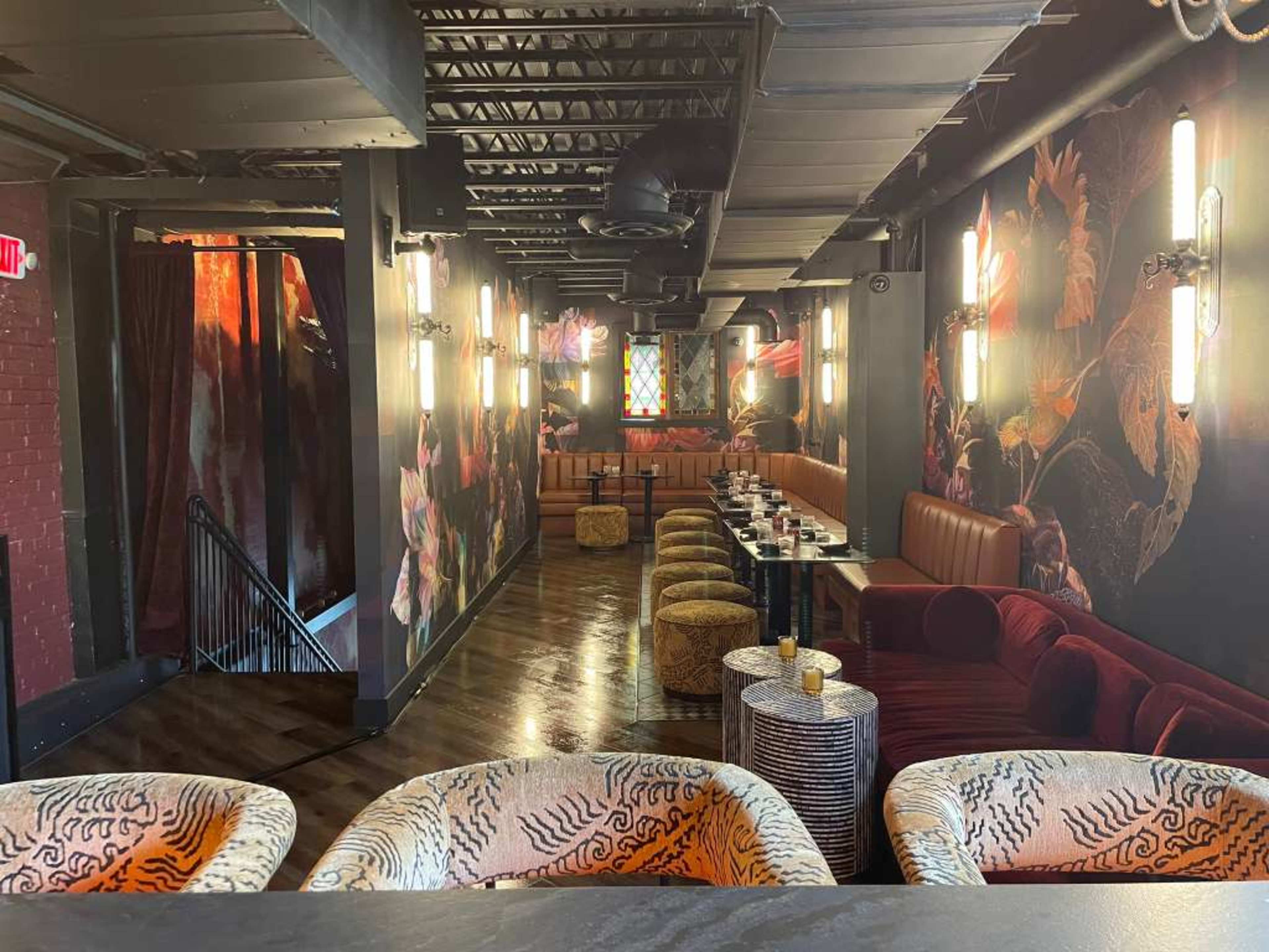 The image shows a dimly lit restaurant interior featuring plush seating, a long table set for dining, and decorative walls with vivid artwork.