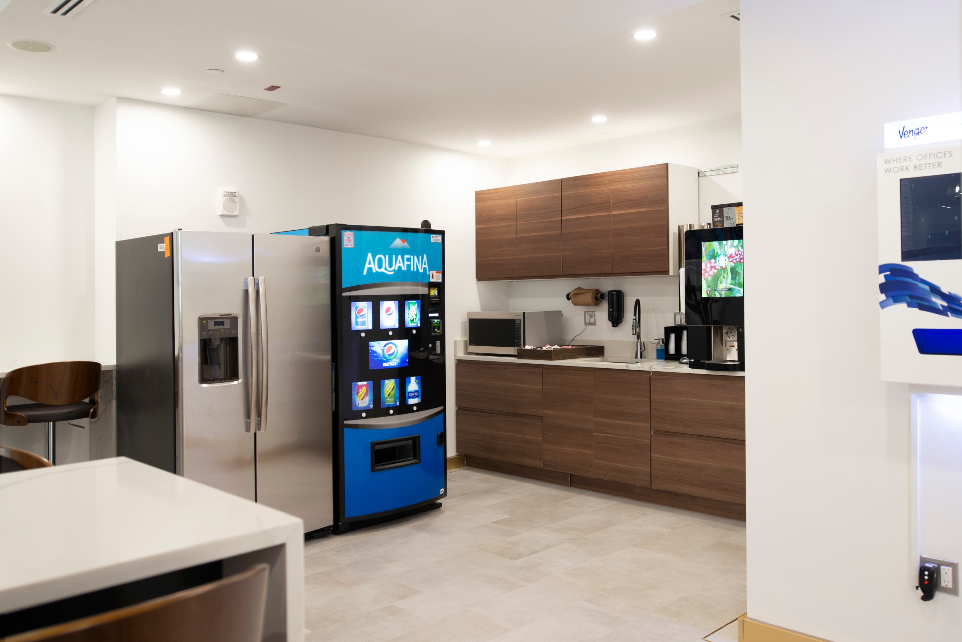 The image shows a modern office kitchenette with a stainless steel refrigerator, a wooden cabinet, and a blue Aquafina vending machine.