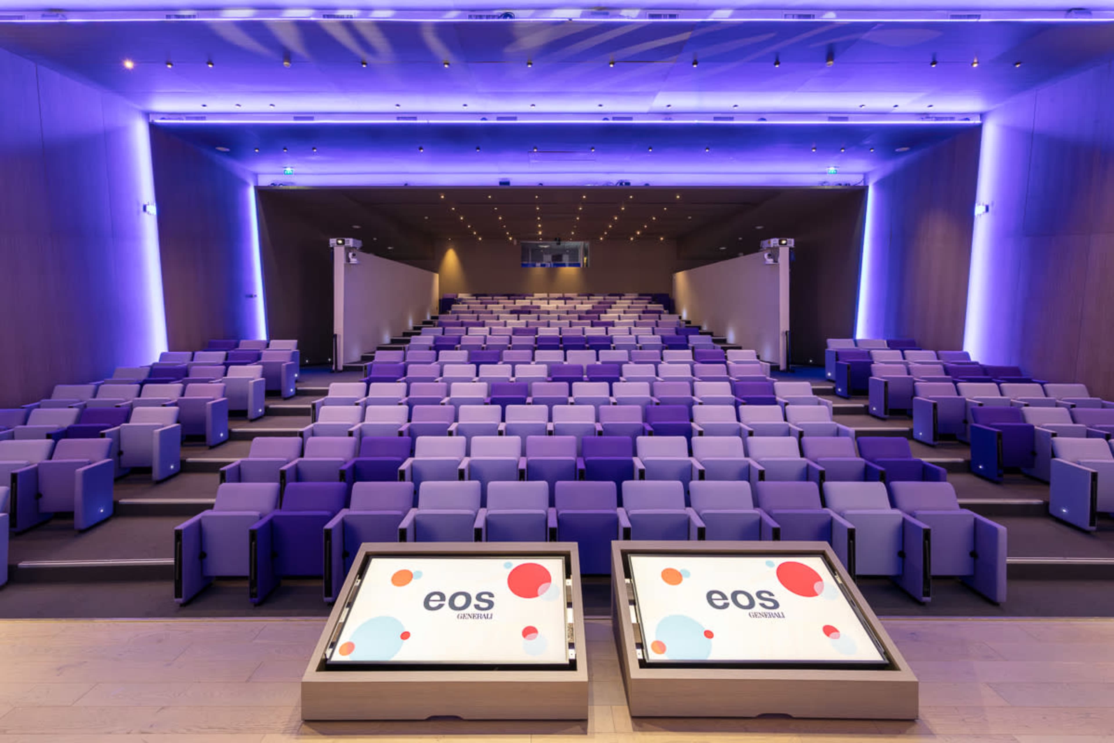 The image shows a modern auditorium with purple seating arranged in a tiered layout, illuminated by ambient lighting along the ceiling.