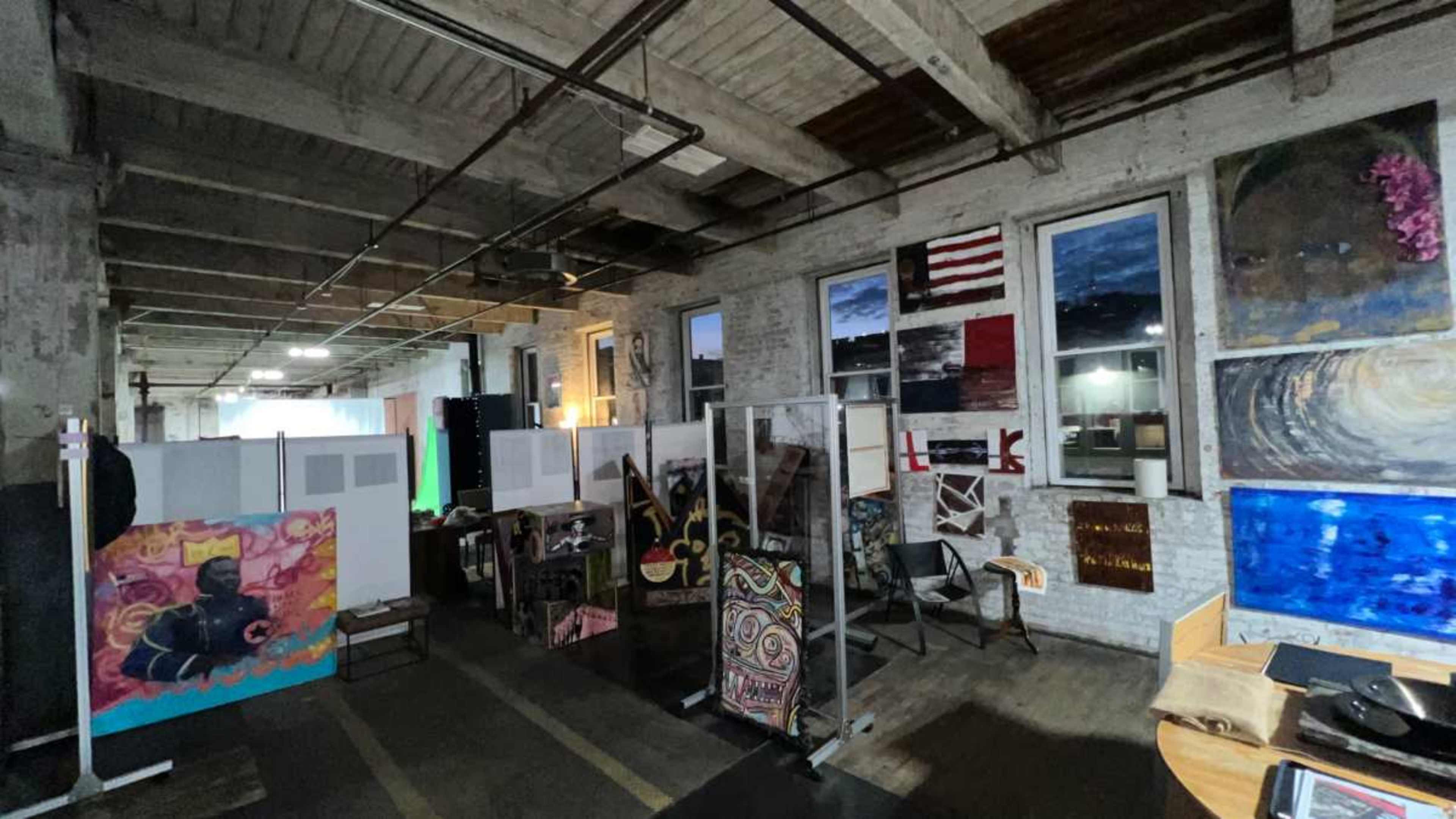 The image shows an industrial-style room filled with various artwork displayed on walls and partition screens.