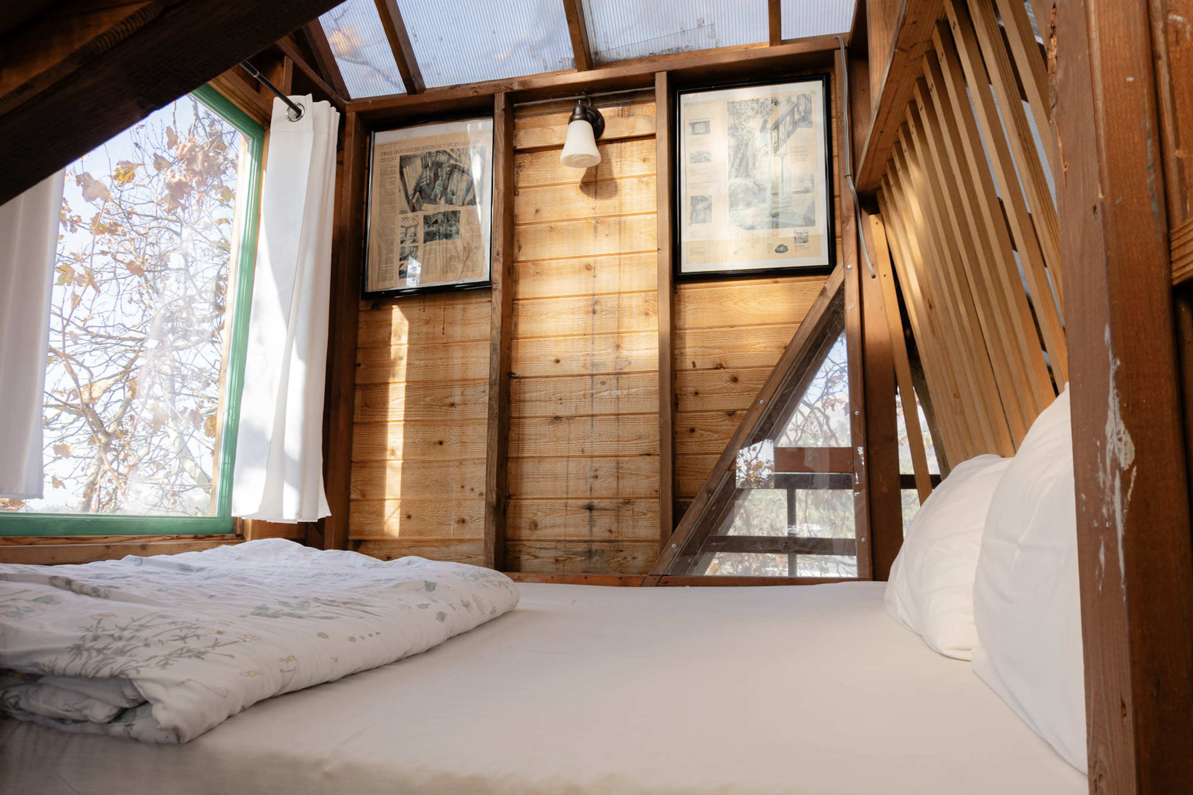 The image shows a wooden loft bedroom featuring a bed with white bedding, framed artwork on the walls, and large windows allowing natural light to enter.