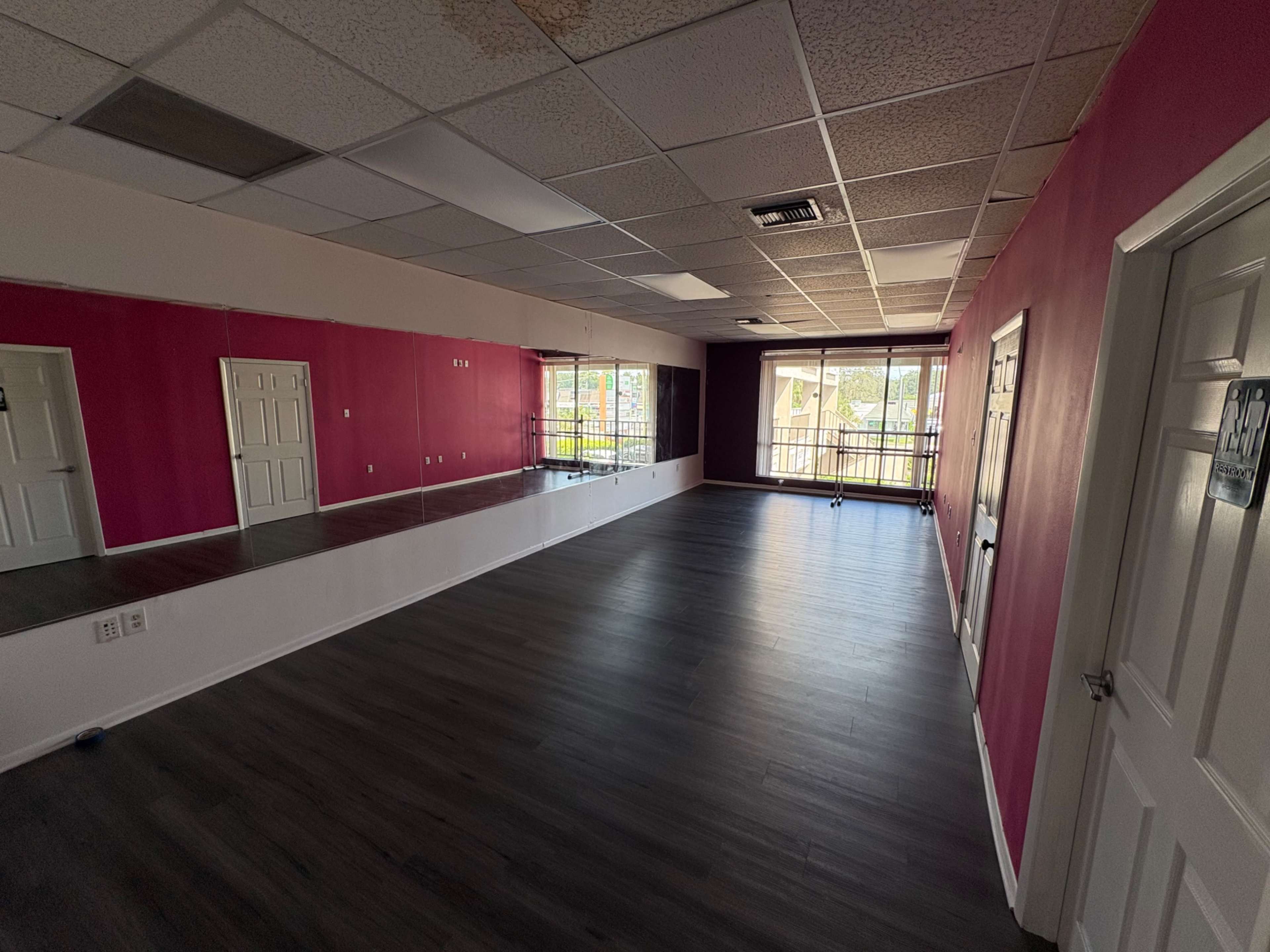 The image shows a spacious room with pink walls, a mirrored wall, and large windows allowing natural light to enter.