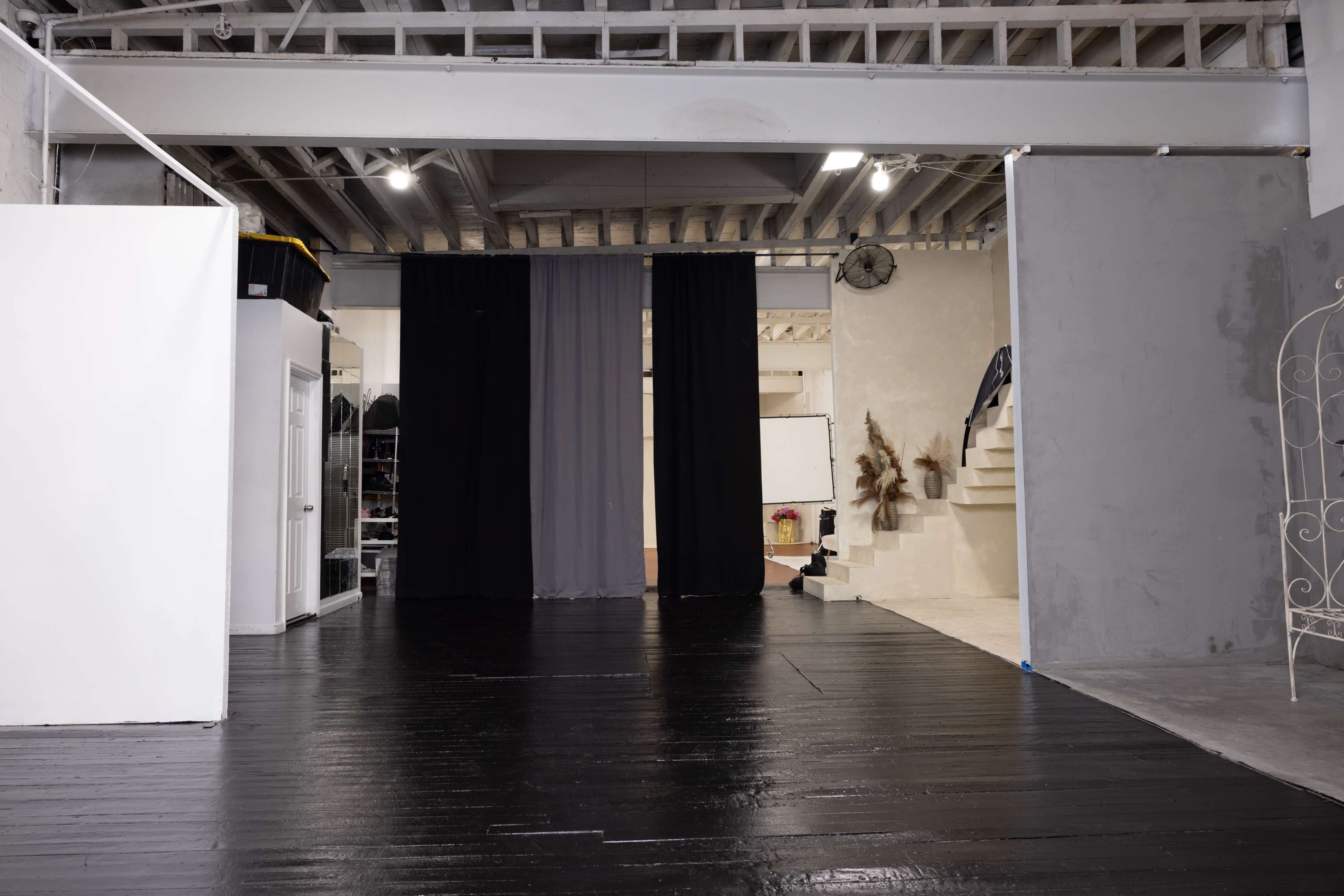 The image shows a spacious studio with dark wooden floors, a black curtain backdrop, and stairs leading to an upper level.
