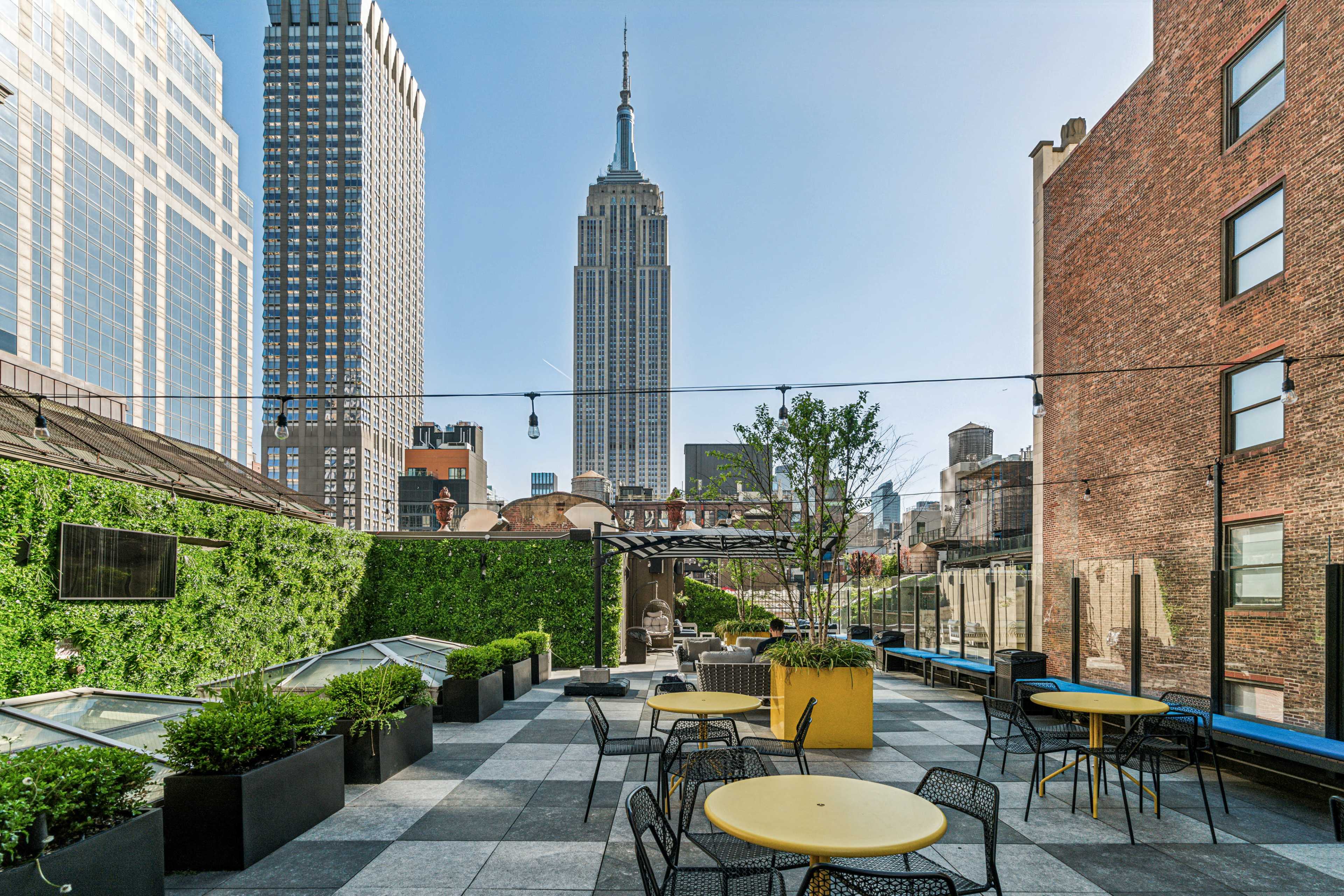 The image shows a rooftop terrace with circular tables and chairs, greenery along the walls, and the Empire State Building prominently visible in the background.