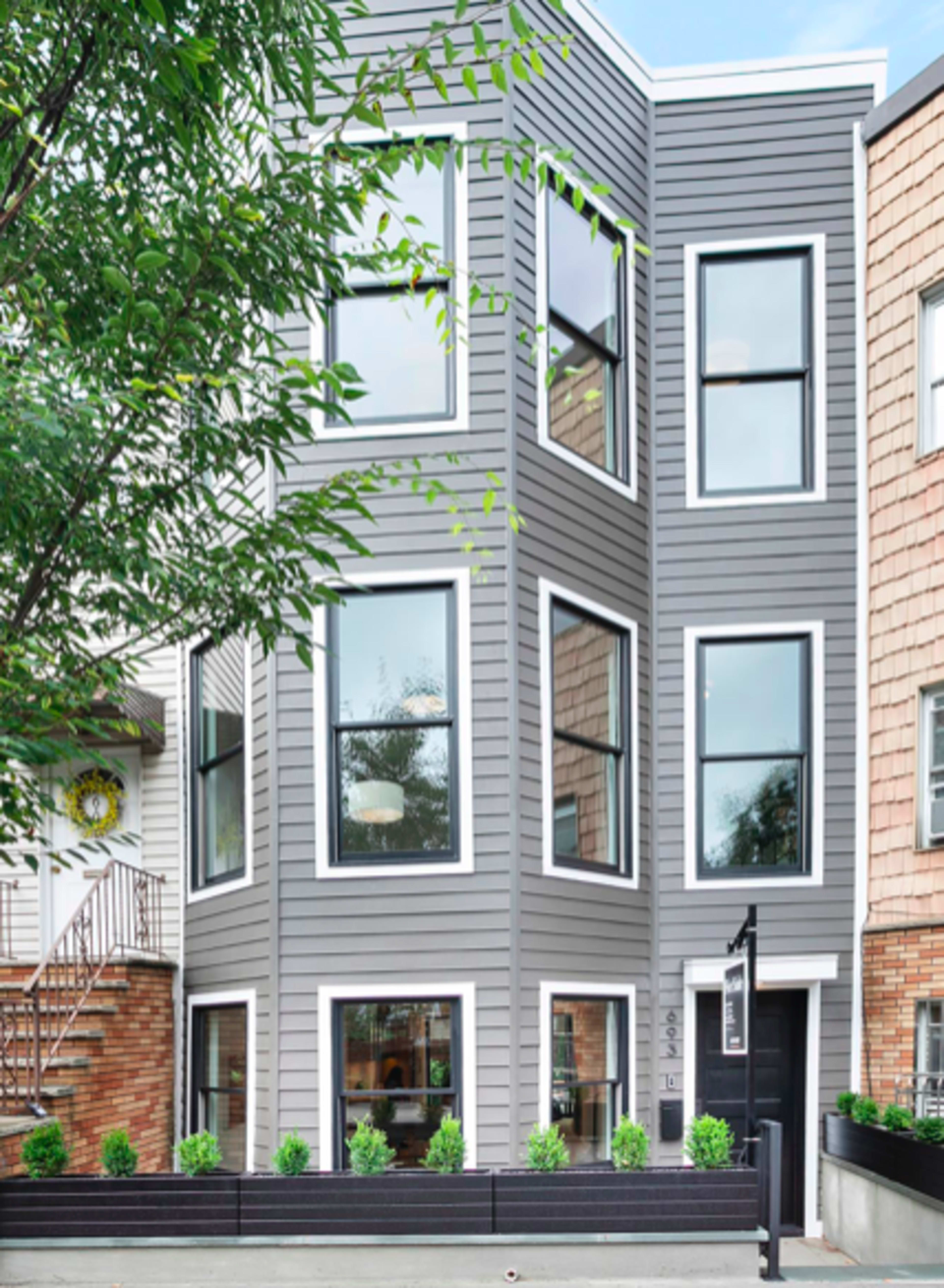 A modern gray house with large windows and a sleek black front door, situated next to neighboring houses with different architectural styles.