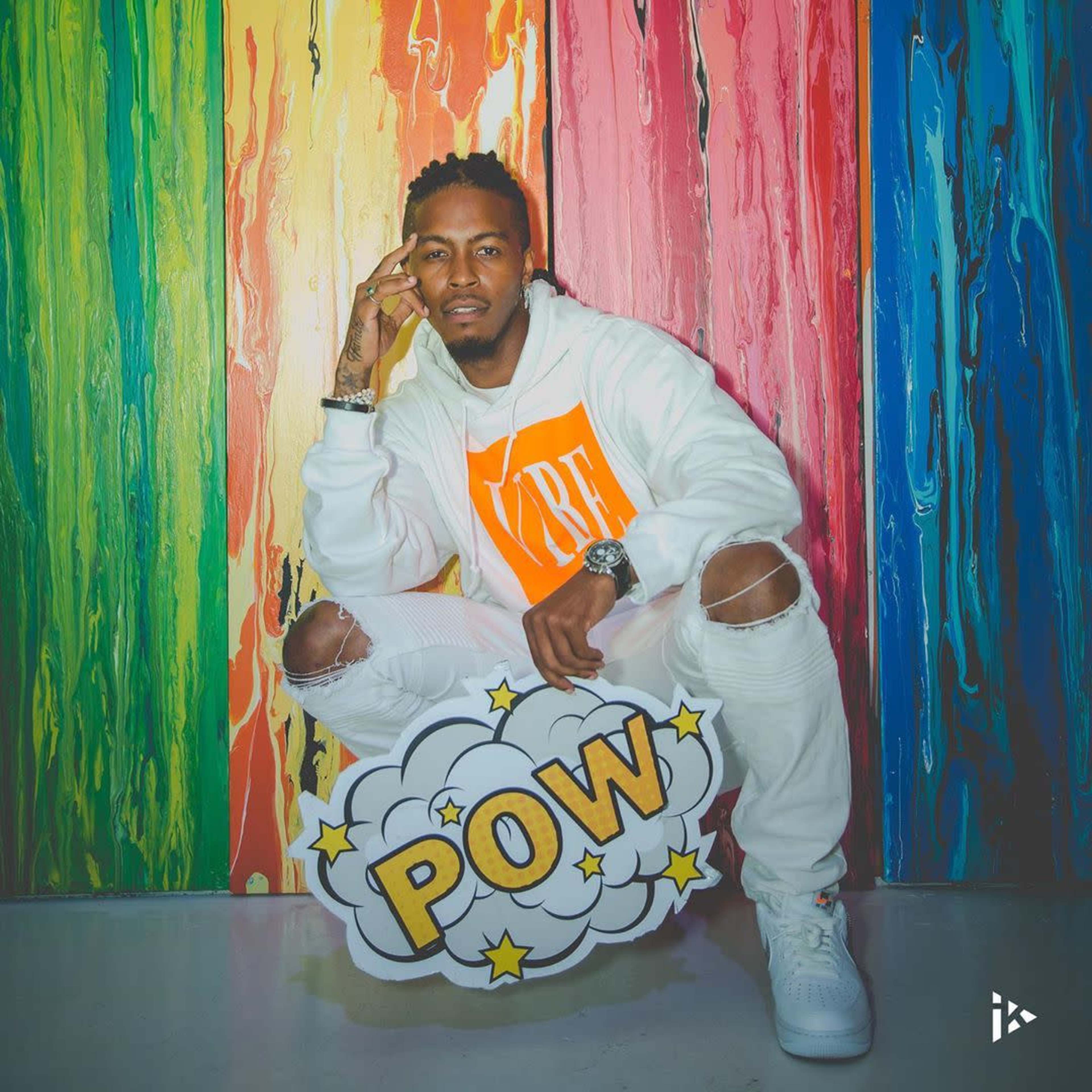 A man in a white outfit poses with a stylized "POW" graphic in front of a colorful, striped background.